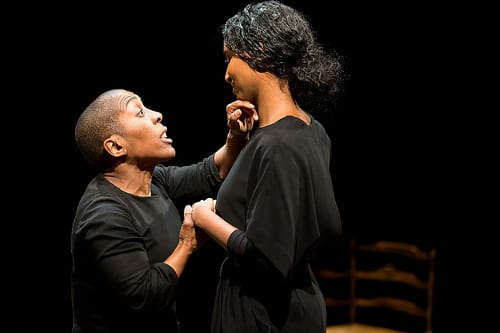 Two performers wearing black dresses hold each other, one wipes something off of the others chin lovingly.