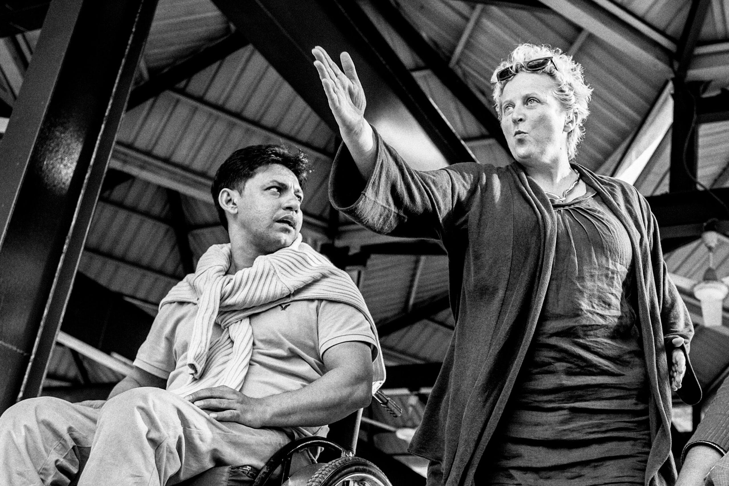 A black and white photo of Jenny Sealy directing. She is standing with a performer who is using a wheelchair. Jenny is pointing to something out of frame.