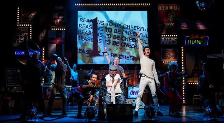 All performers on a cluttered and busy stage, with their arms outstretched to the air. The text behind them reads Reasons to be Cheerful.