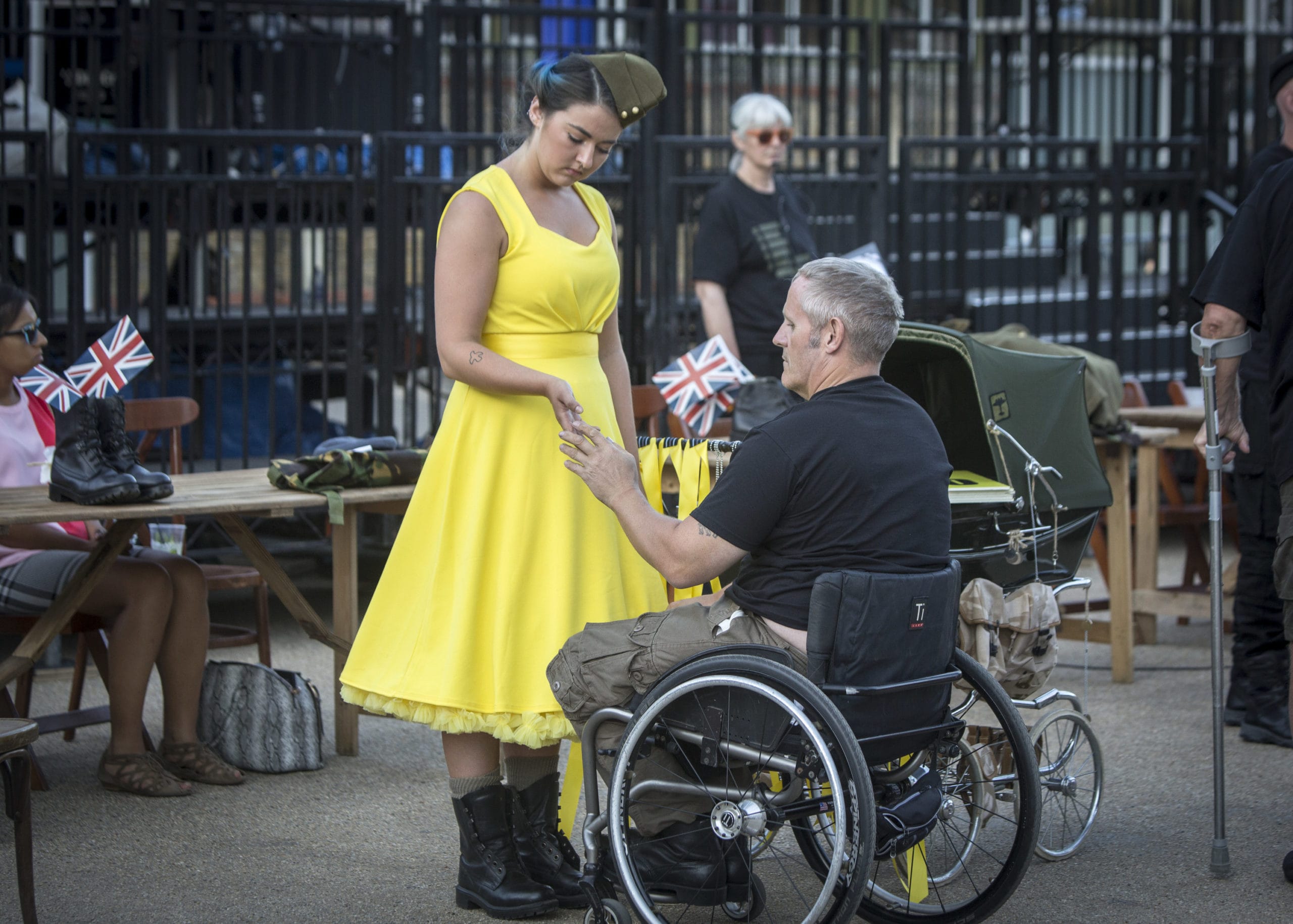 A man in a wheelchair grasps the hand of a young woman, stood up, wearing a vibrant yellow dress.