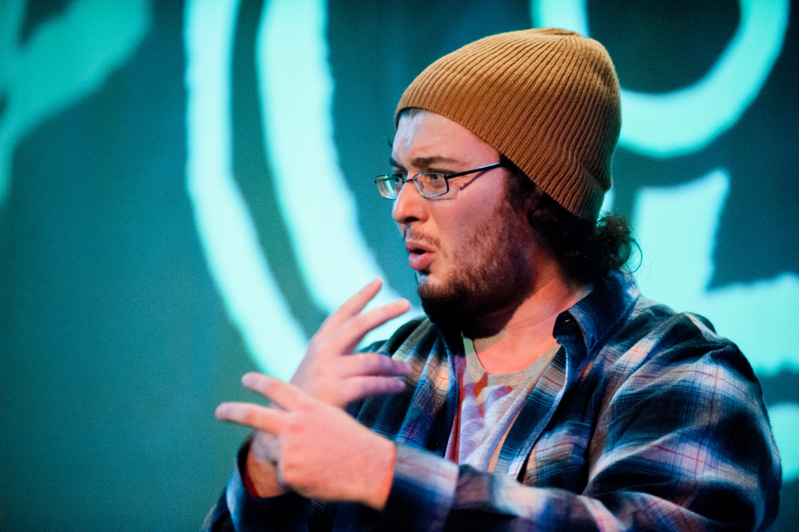 A man wearing a mustard beanie and blue flannel shirt signs to an audience. He has a brown beard and glasses.