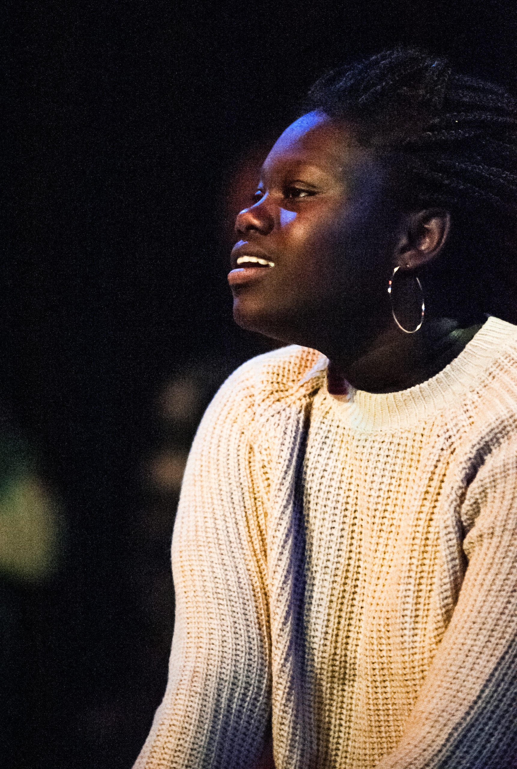 A young Black woman wearing a white knitted jumper looks away from the camera and smiles.