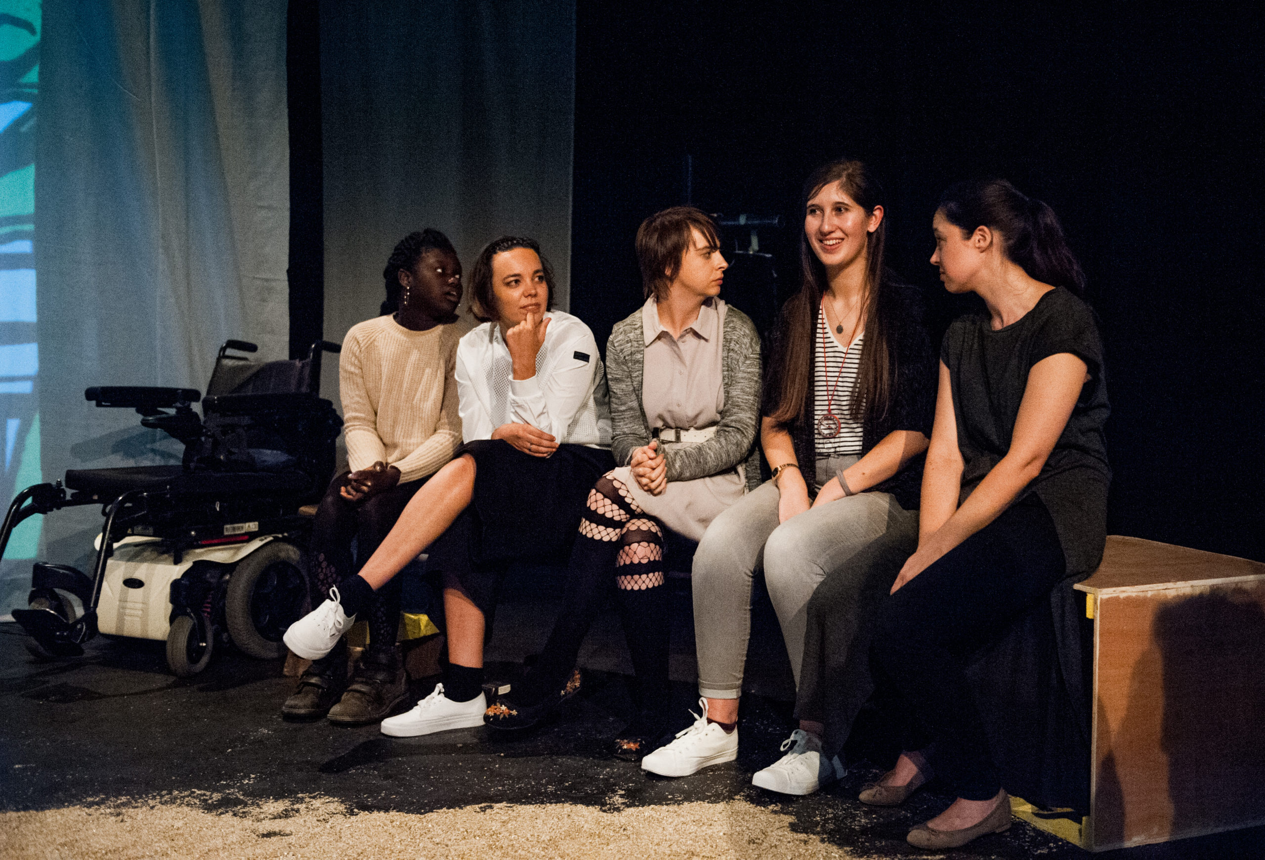 Five performers sit in a row, most with their legs crossed.
