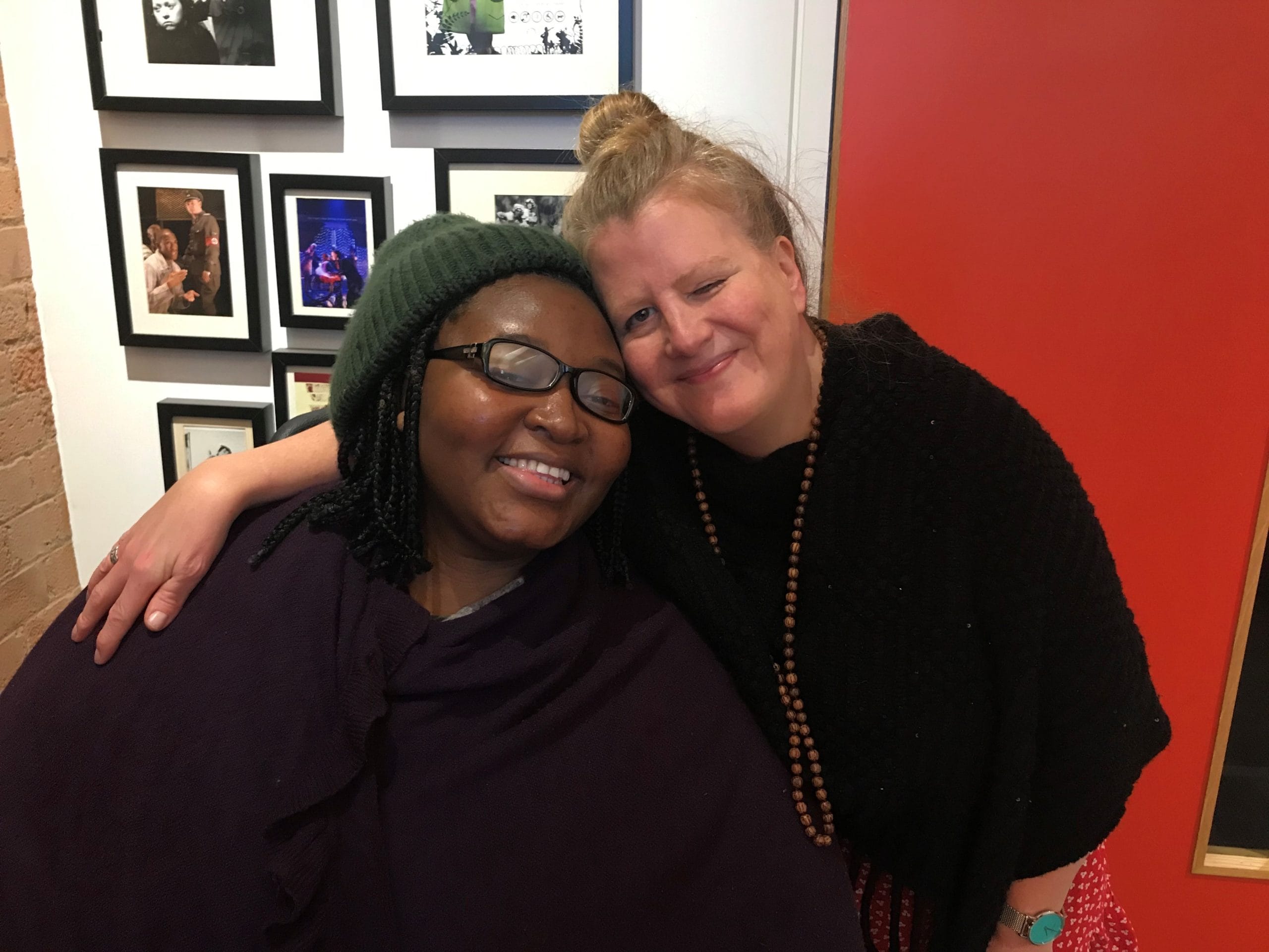 Matilda, a black woman, with short, thick braids, wears a green hat, sits next to Jodi, a white woman, with blonde hair who has her arm around Matilda. They are bth smiling at the camera.