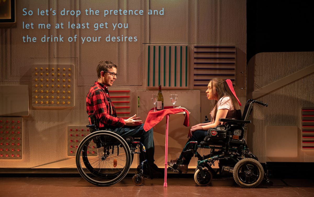 Two performers in wheelchairs sit opposite each other, at a table set up for a dinner date.