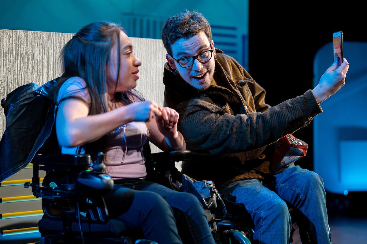 Two performers in wheelchairs sit next to each other; the man leans into the woman holding a phone up to take a selfie.