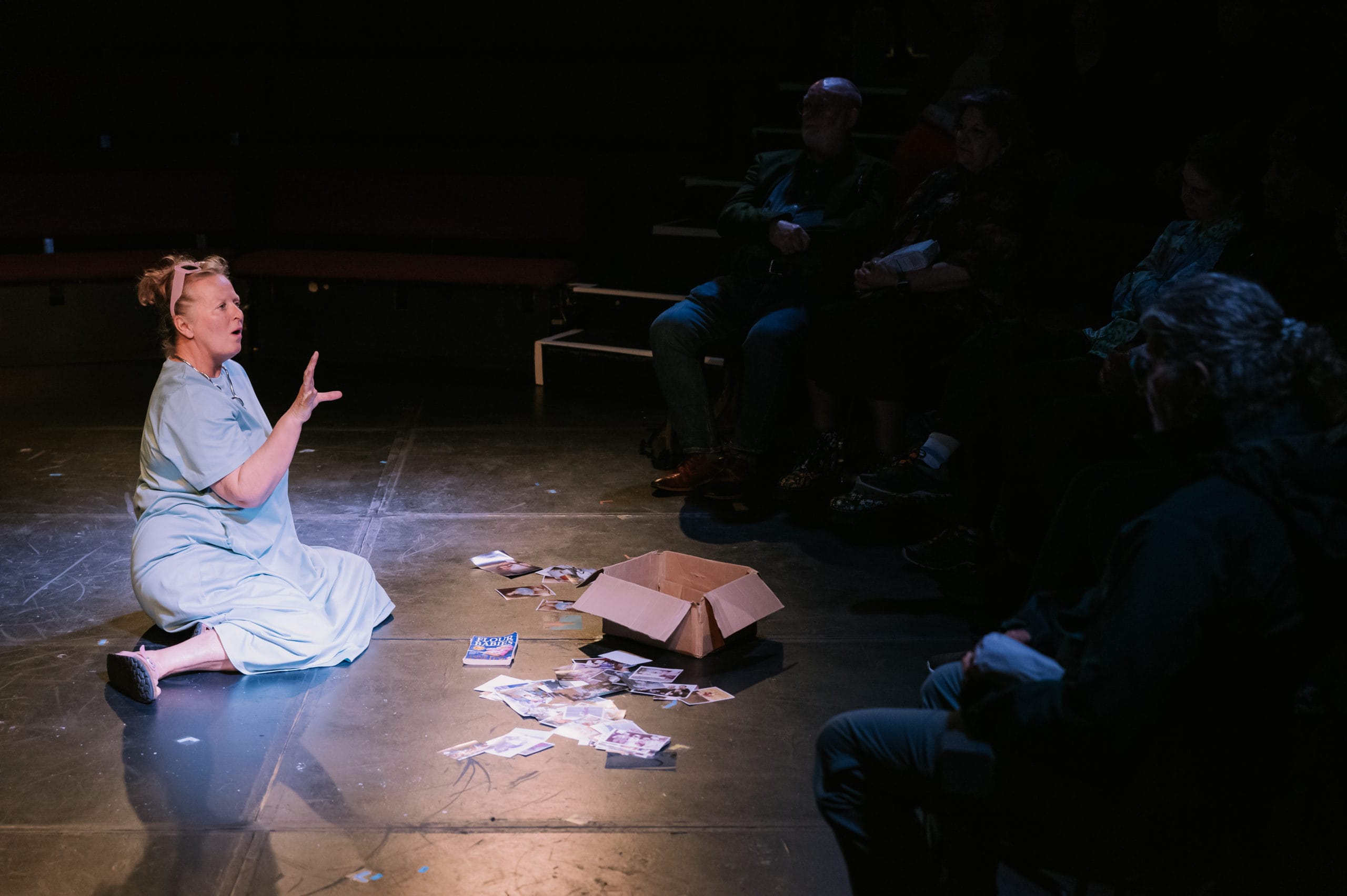 A woman sits alone in a spotlight, in front of her is a box of photographs spilled all over the floor.