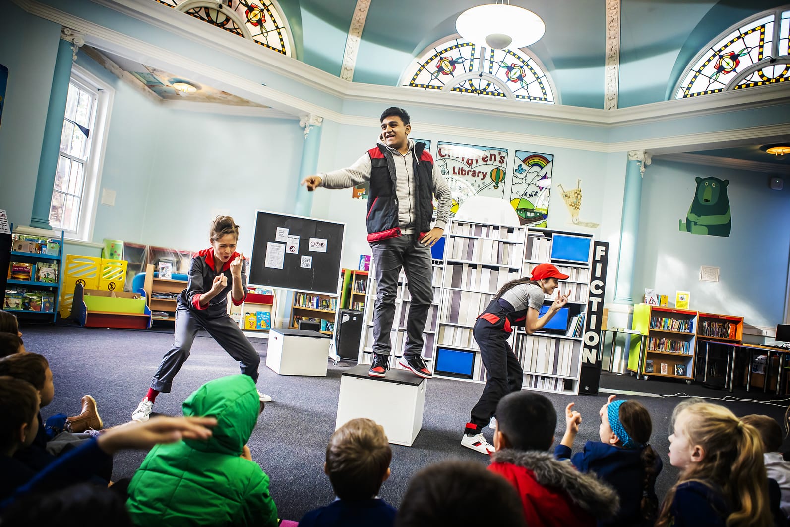A group of young actors perform to a group of children in a library.