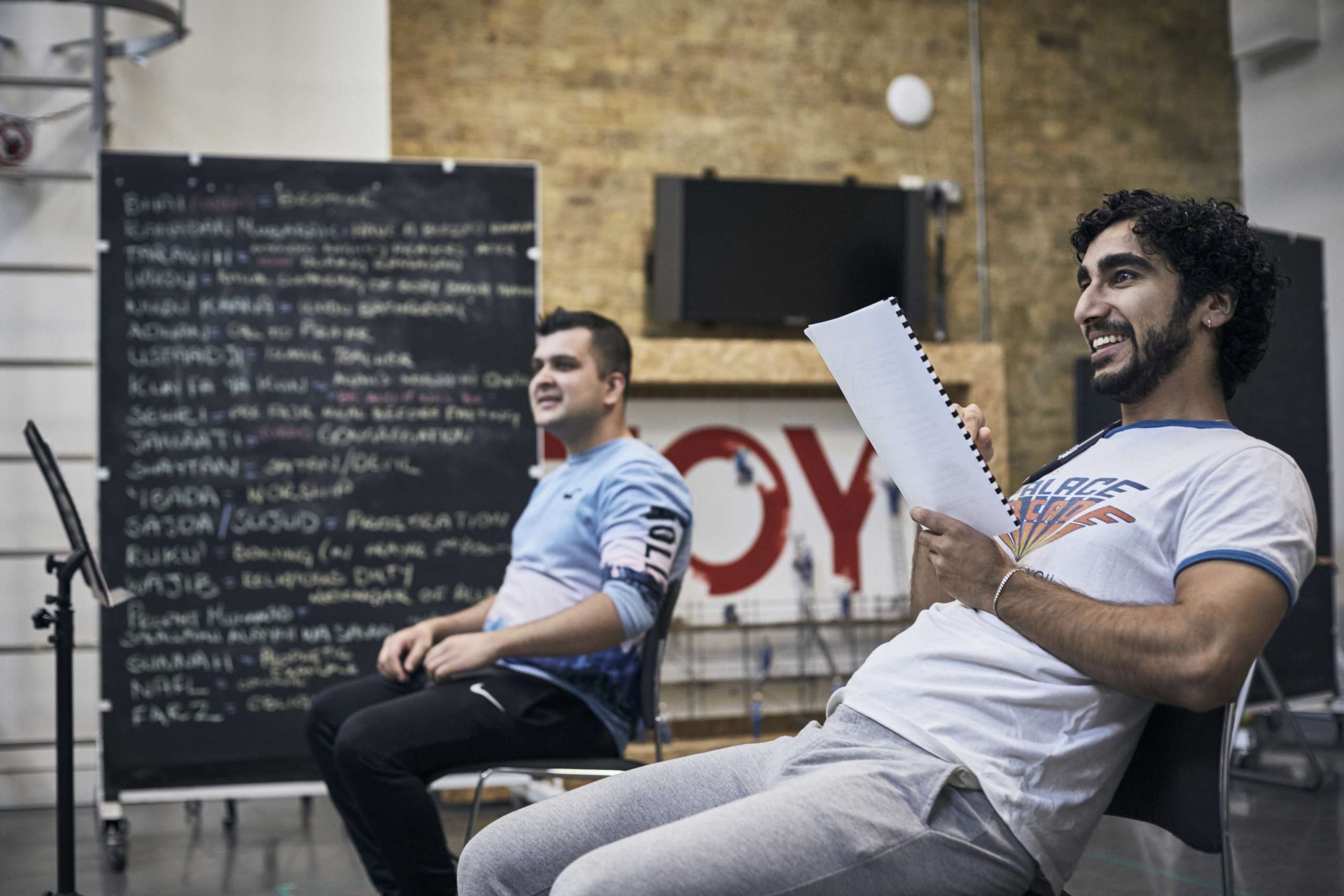 Two men sit in a rehearsal room, holind scripts.