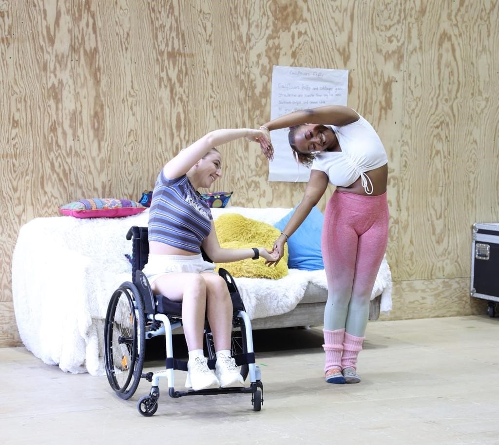 A wheelchair user and a woman standing next to her form a heart shape with their arms