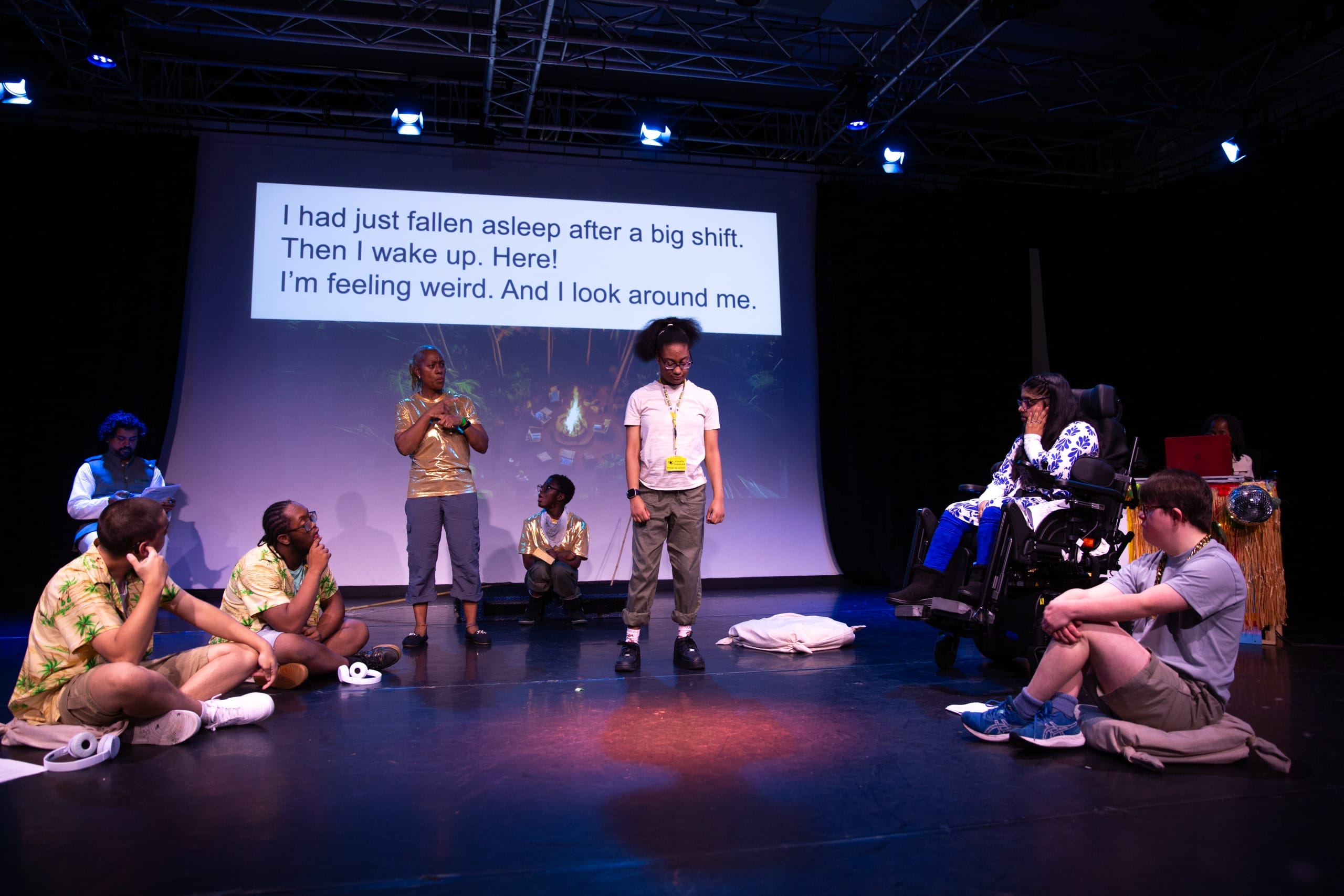 Production photo from The Anchors and The Sails. Several performers in a semicircle onstage. One actress is standing and one interpreter is standing, the rest are seated. Captions are projected on the wall behind the performers.