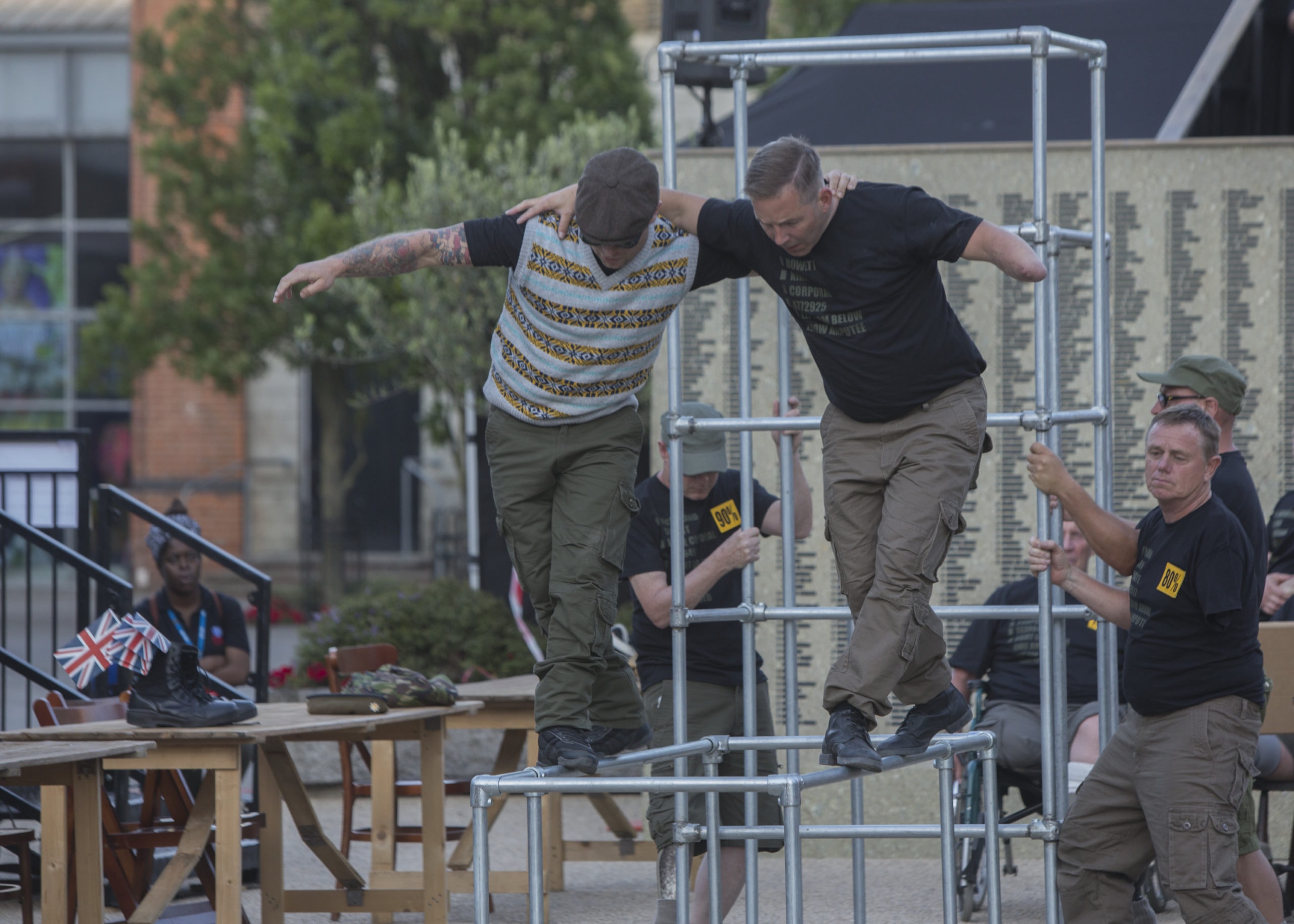 Two performers help each other balanc along a metal scaffolding, with their arms around each others shoulders