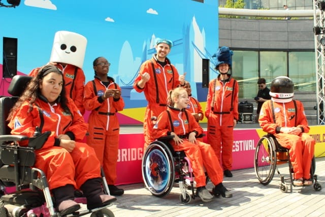 A series of participants of the children's drama group for 6-11 year olds, all dressed in orange boiler suits outside, with three sitting in wheelchairs.
