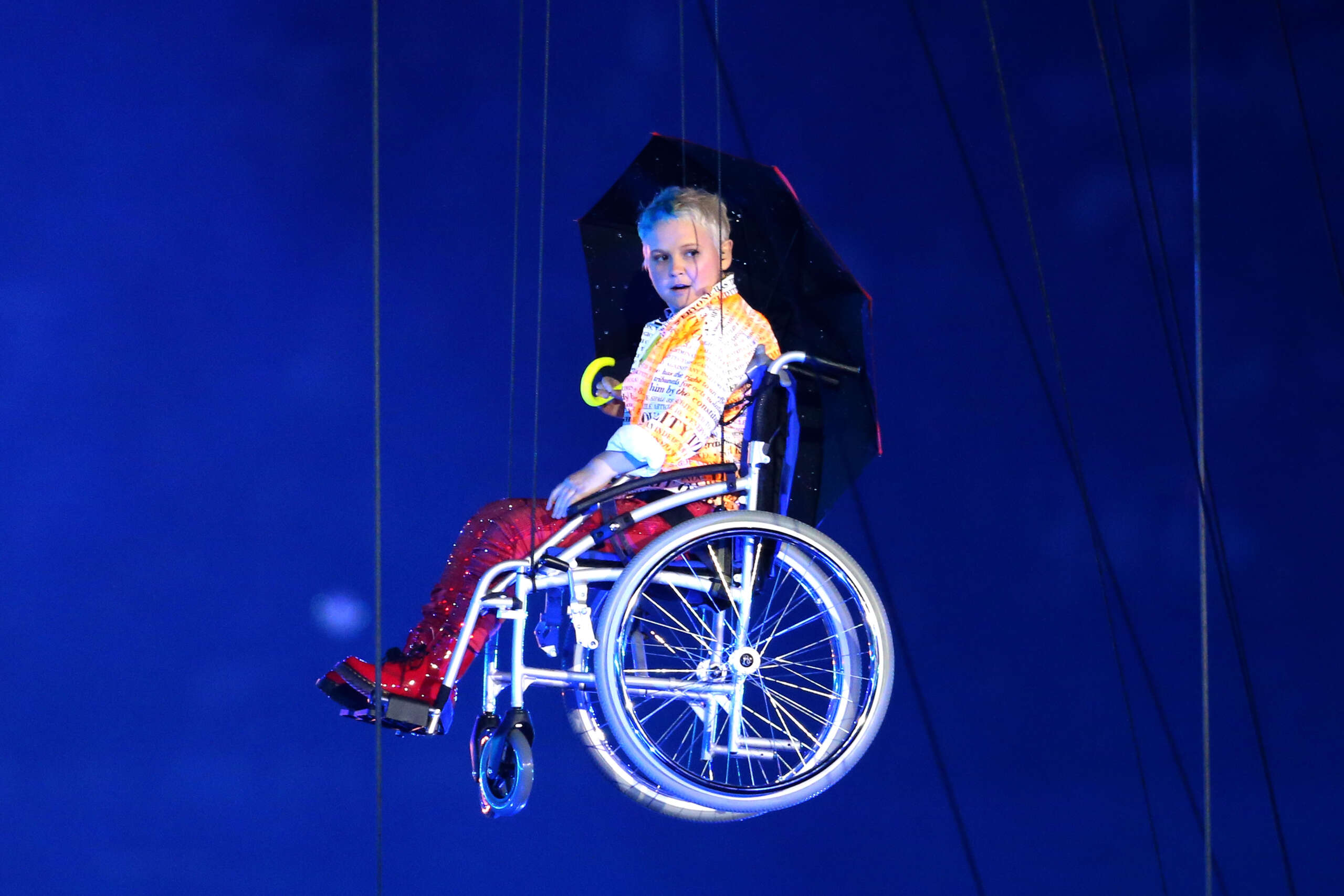 A young performer in a wheelchair, is suspended in the air. He is white, with short blonde hair, wearing a yellow shirt, red checked trousers, and red boots. He is holding a large black umbrella.