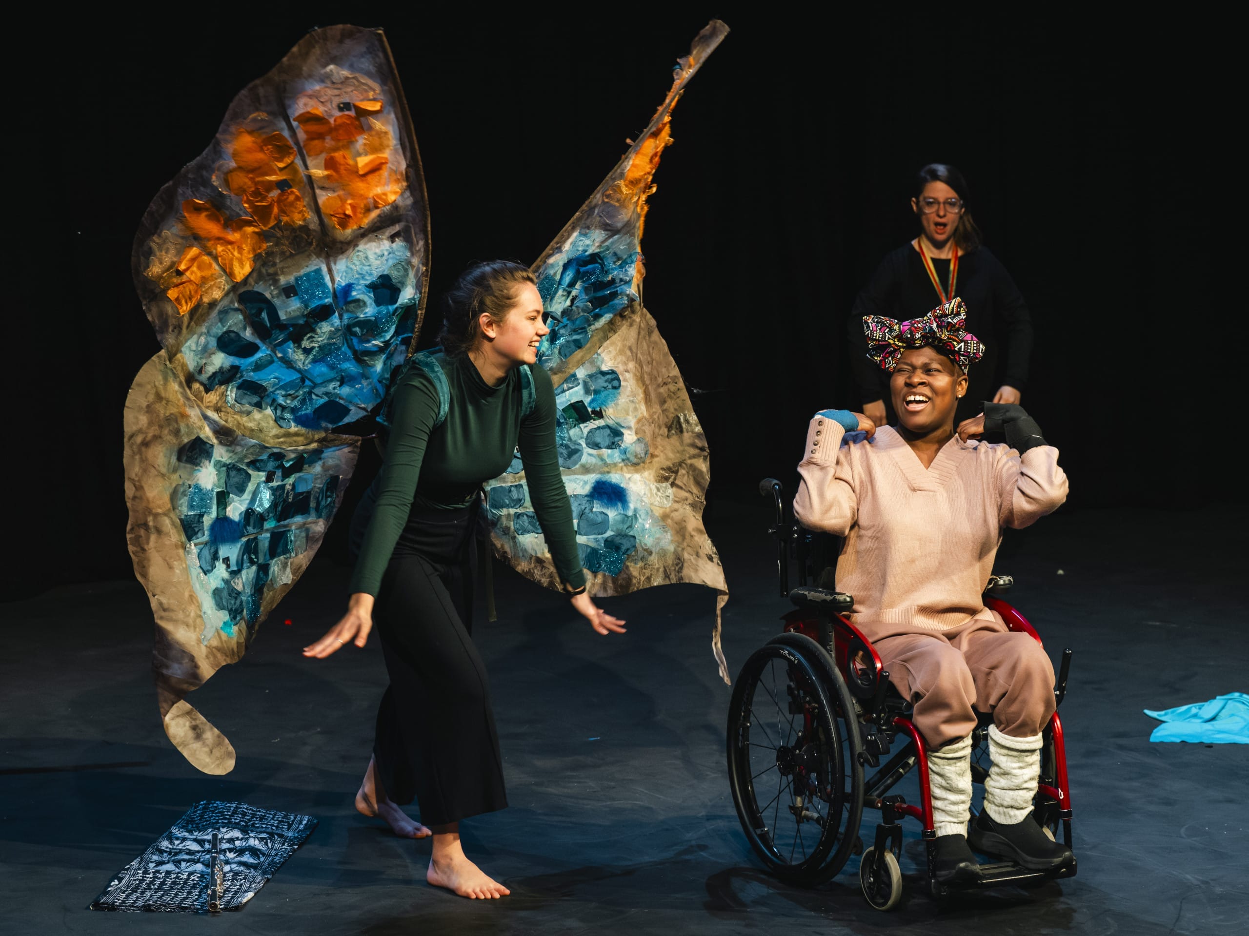 A wheelchair user wearing all beige sits besides a woman dressed in a butterfly costume, wearing large butterly wings in blue and orange. They smile at each other.