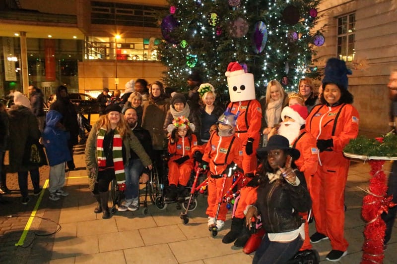 A group of children and adults stand together in front of a huge christmas tree. Some of them are dressed up in orange astronaut costumes. Some of them are using wheelchairs or walking aids.
