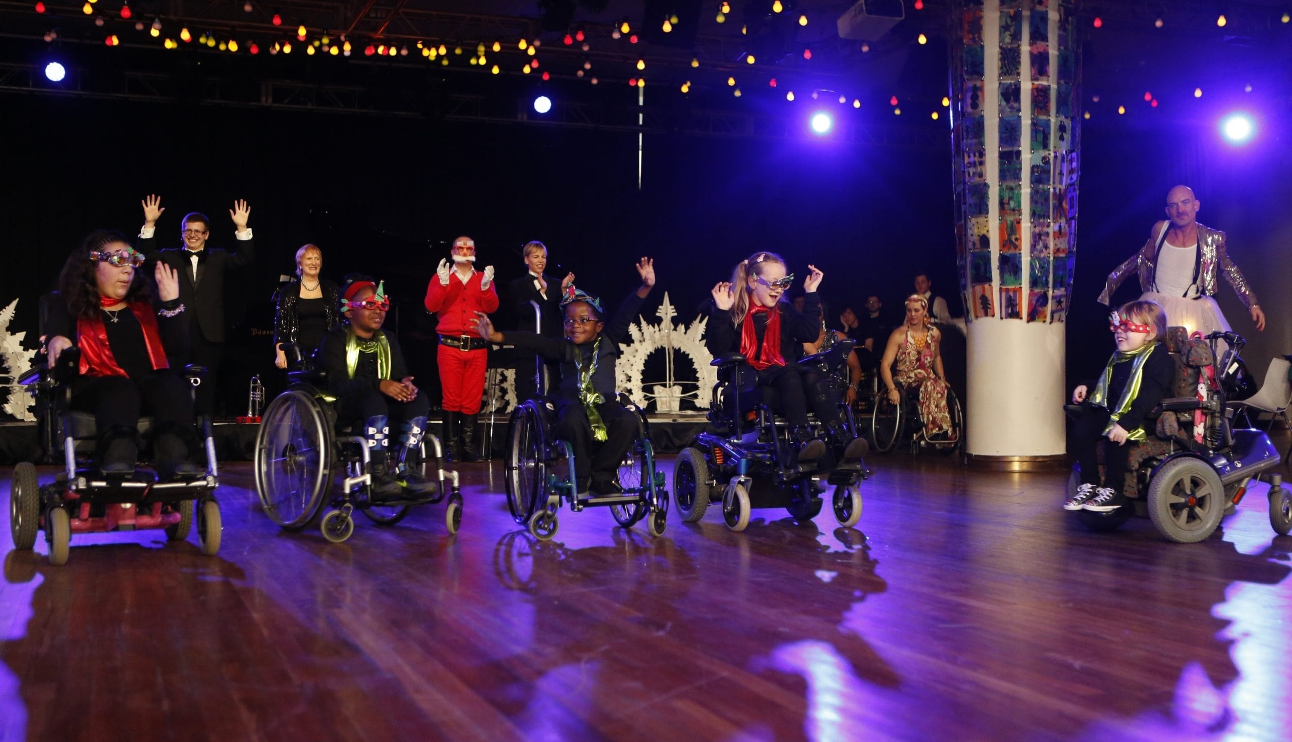 A group of young children in wheelchairs are in the middle of the performance space, they are surrounded by Christmas decorations.