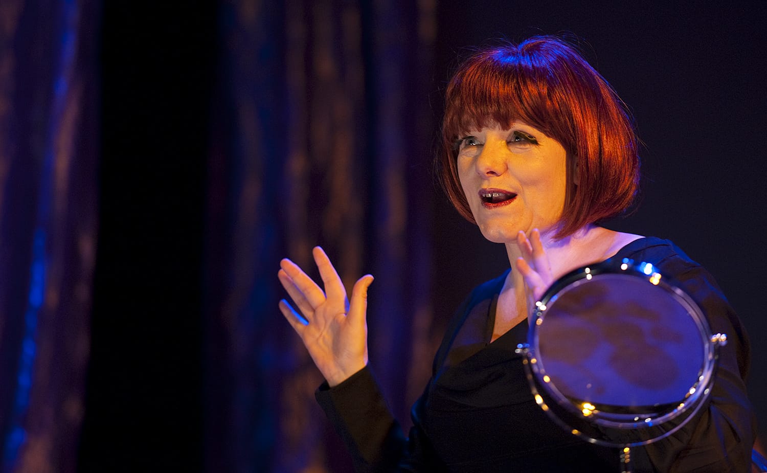 Sue Parker stands with her arms outstretched. With a vibrant red wig on her head.
