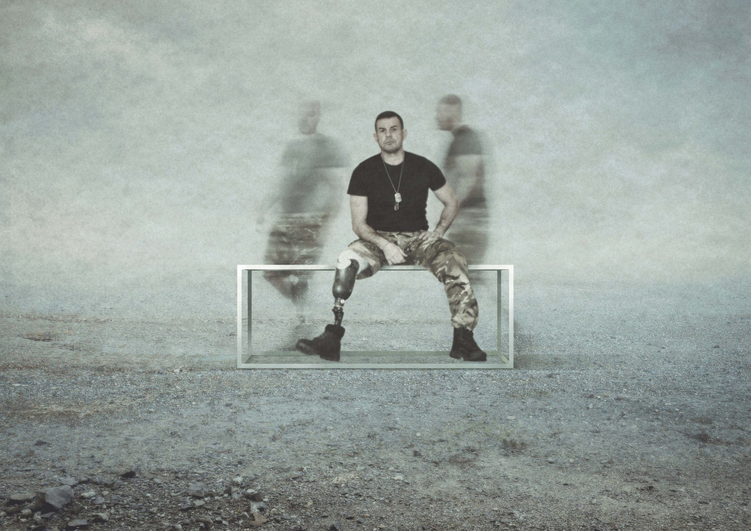 A wounded serviceman with two prosthetic legs sits on a metal structure. The blurred outline of two people rush behind him. His gaze is fixed on the camera in front of him.