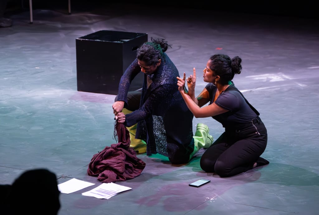 2 woman kneel on the floor wearing black. One signs while the other holds a piece of material tightly.
