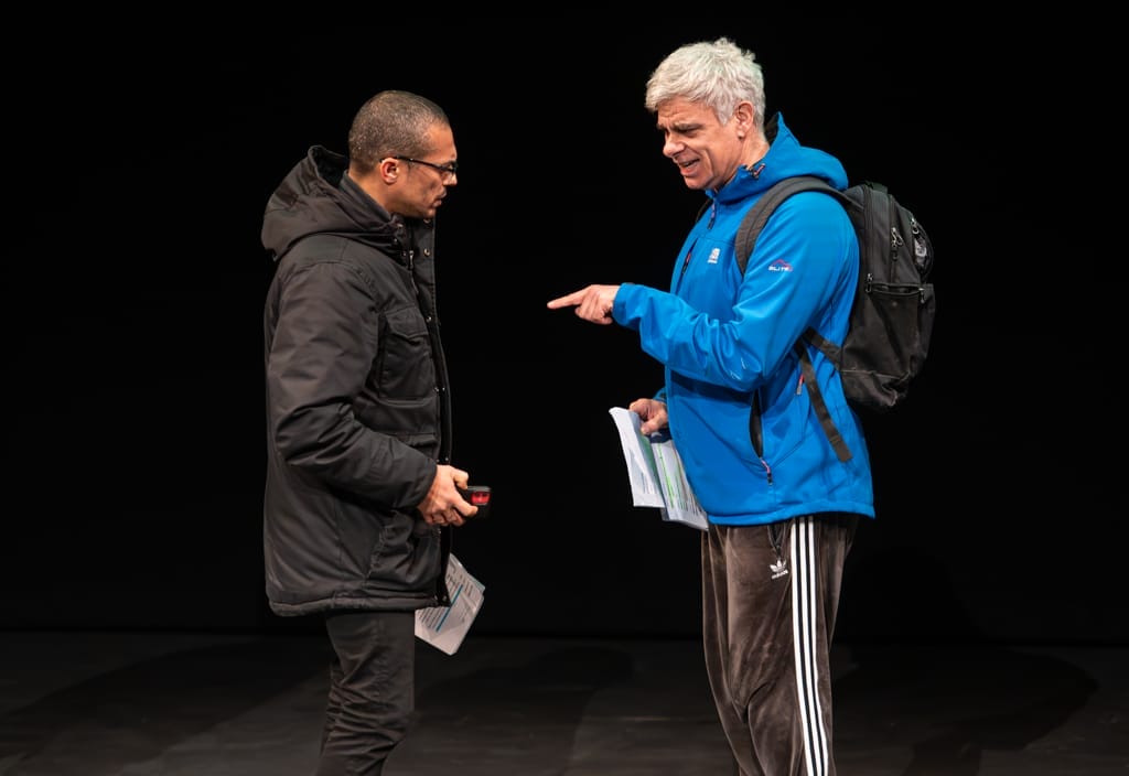 2 actors stand face to face on stage. One wears a blue jacket and a backpack, the other in a black jacket. Both hold scripts in hand.