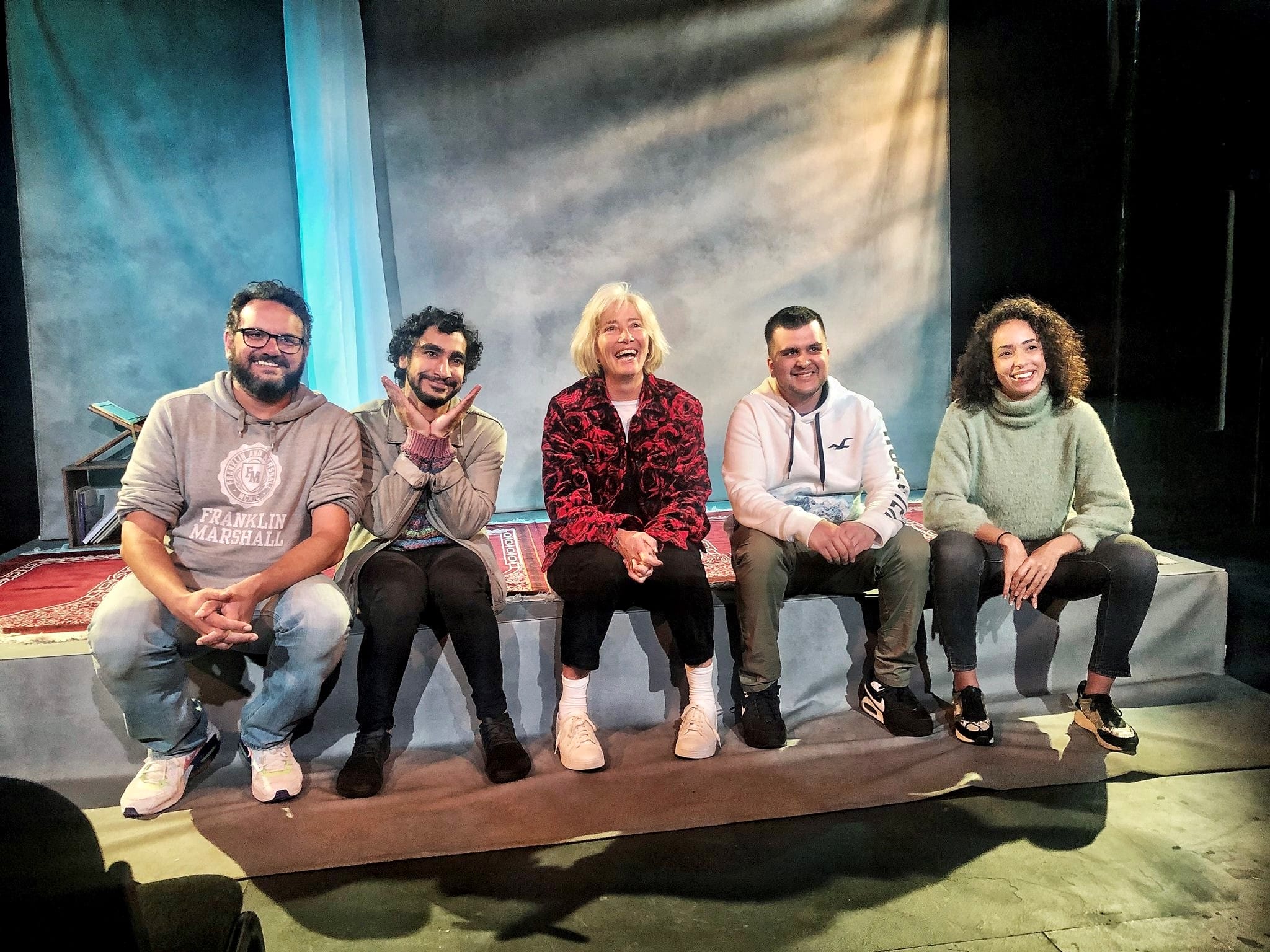 A group of five people sit on the edge of a stage. They are all smiling and laughing at the camera.