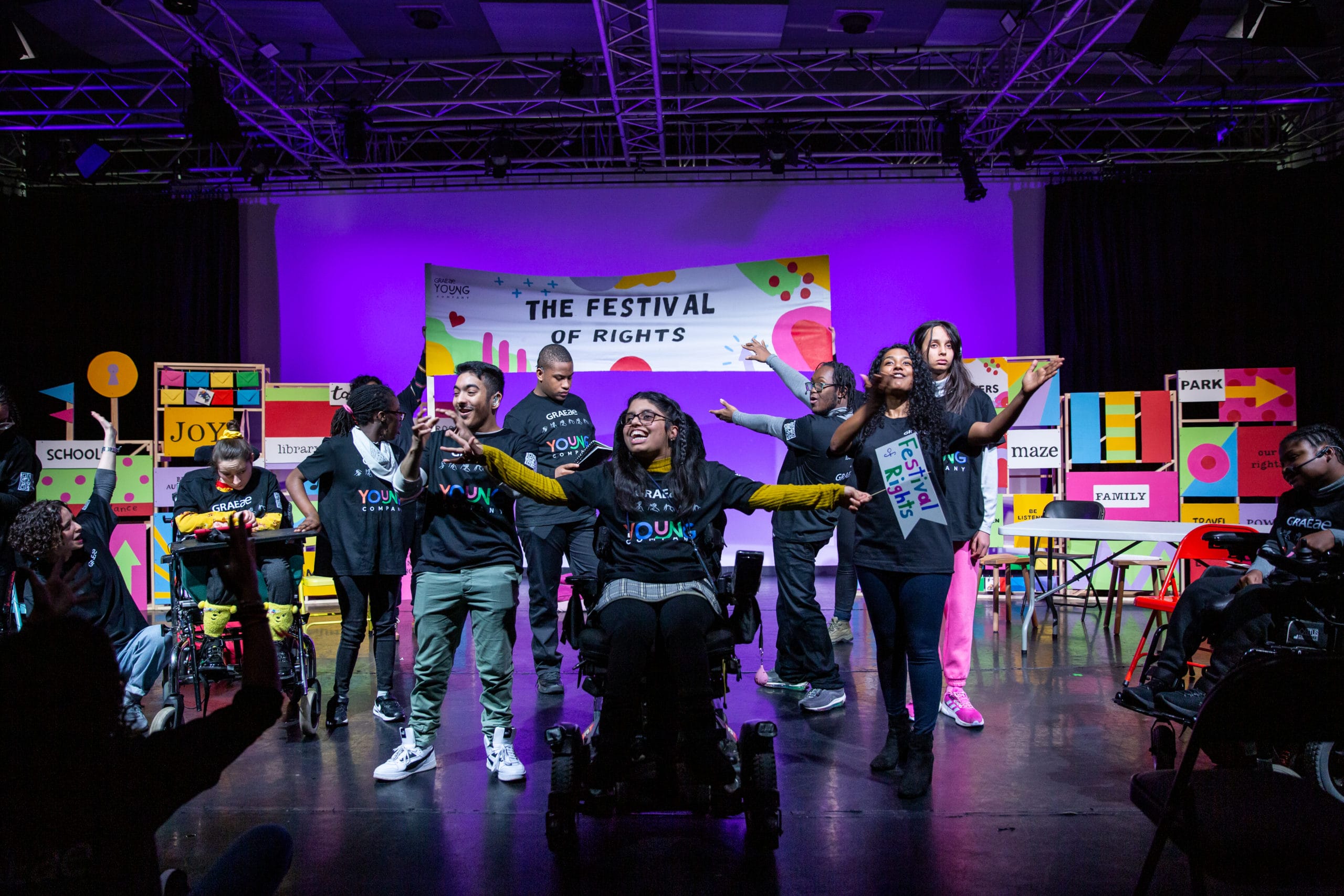 The cast of the Graeae show 'The Festival of Rights' all stand centre stage. A young girl in the middle in a wheelchair has her arms outstretched, and smiles a wide smile.
