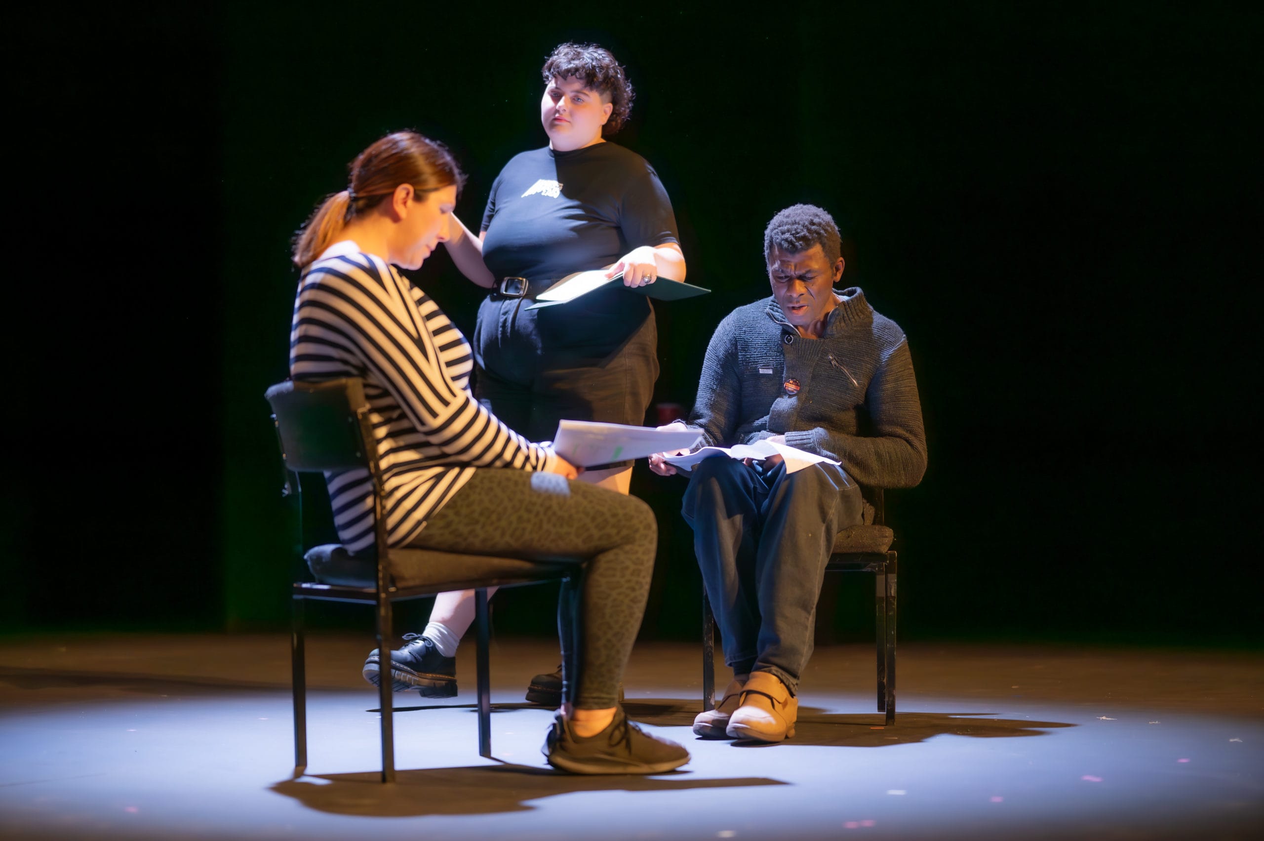 Three performers on stage. Two are sitting in chairs facing each other, and the third is walking behind them. They are all holding scripts.