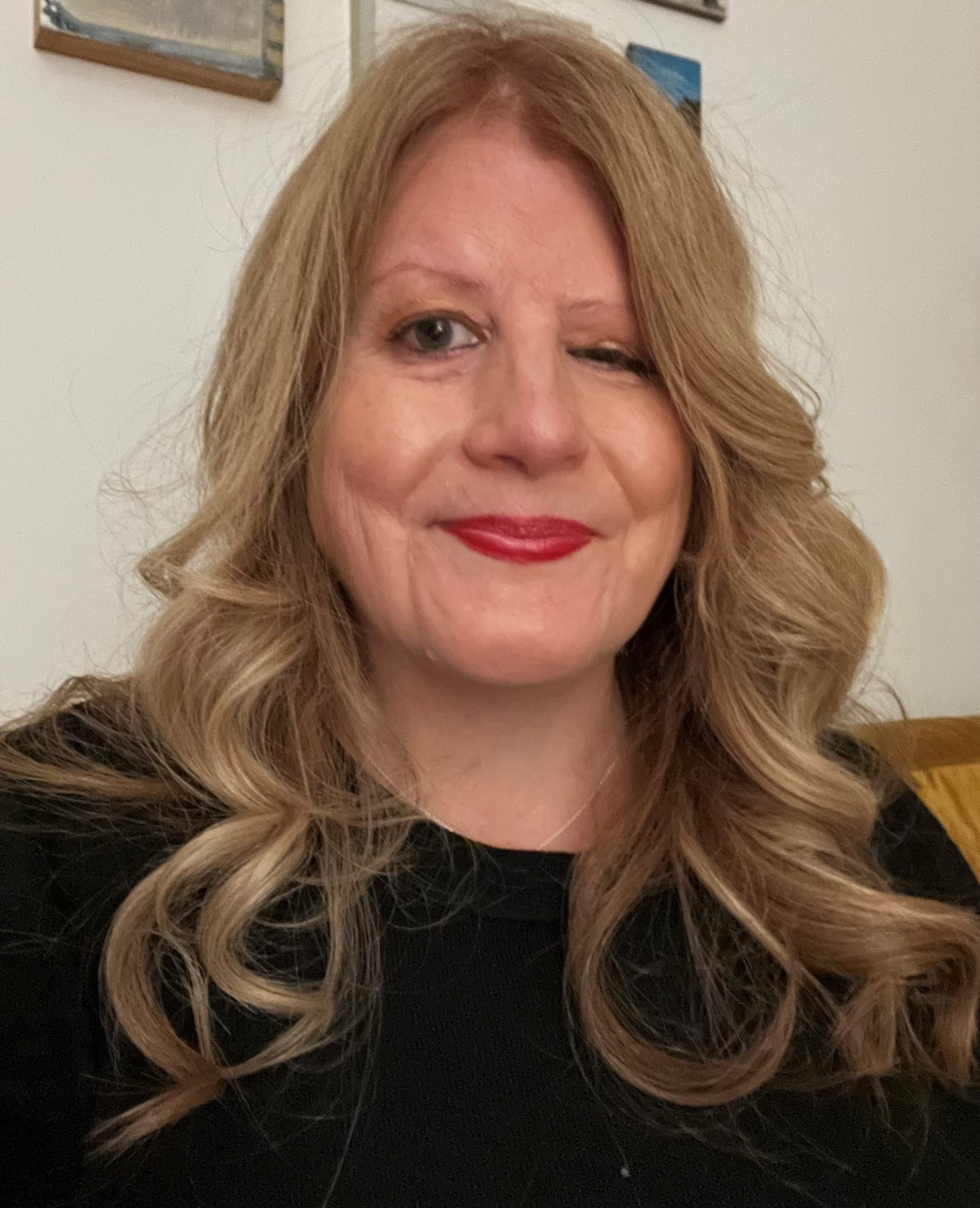 Jodi, a white woman with long blonde hair is stood in front of a plain white backdrop. She is smiling, wearing a black top