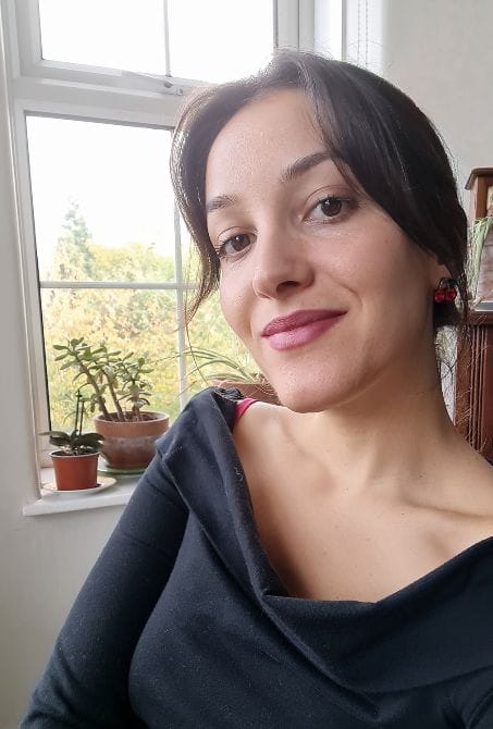 Selfie of a white woman in her early thirties smiling at the camera. She has brown straight hair in a ponytail and wears pink lipstick. There are pots with plants sitting on the windows in the background behind her.