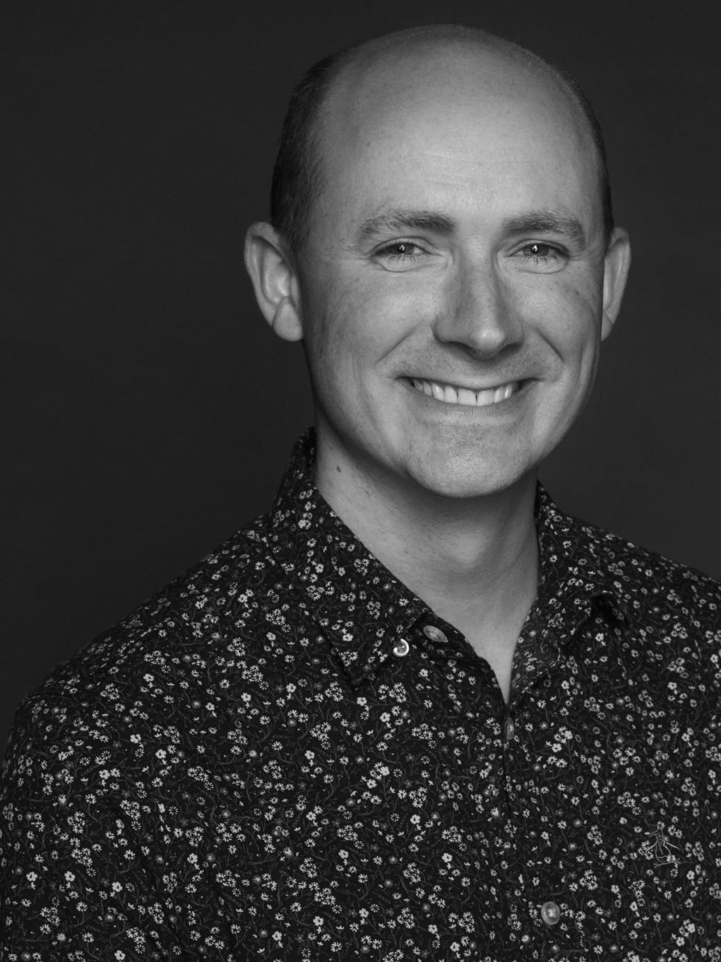 Kevin, a white man, is stood infront of a grey backdrop. The photo is in black and white. He smiles a wide smile, and is wearing a black button-up shirt with small white flowers detailed on it.