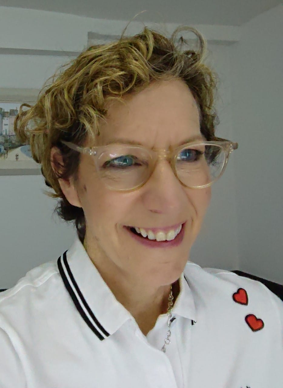 A white woman in their 50’s with short curly blonde coloured hair. Wearing a white polo shirt with black stripes along the collar and 2 red hearts sewn on the shoulder. Laura wears clear framed glasses and smiles away from the camera.