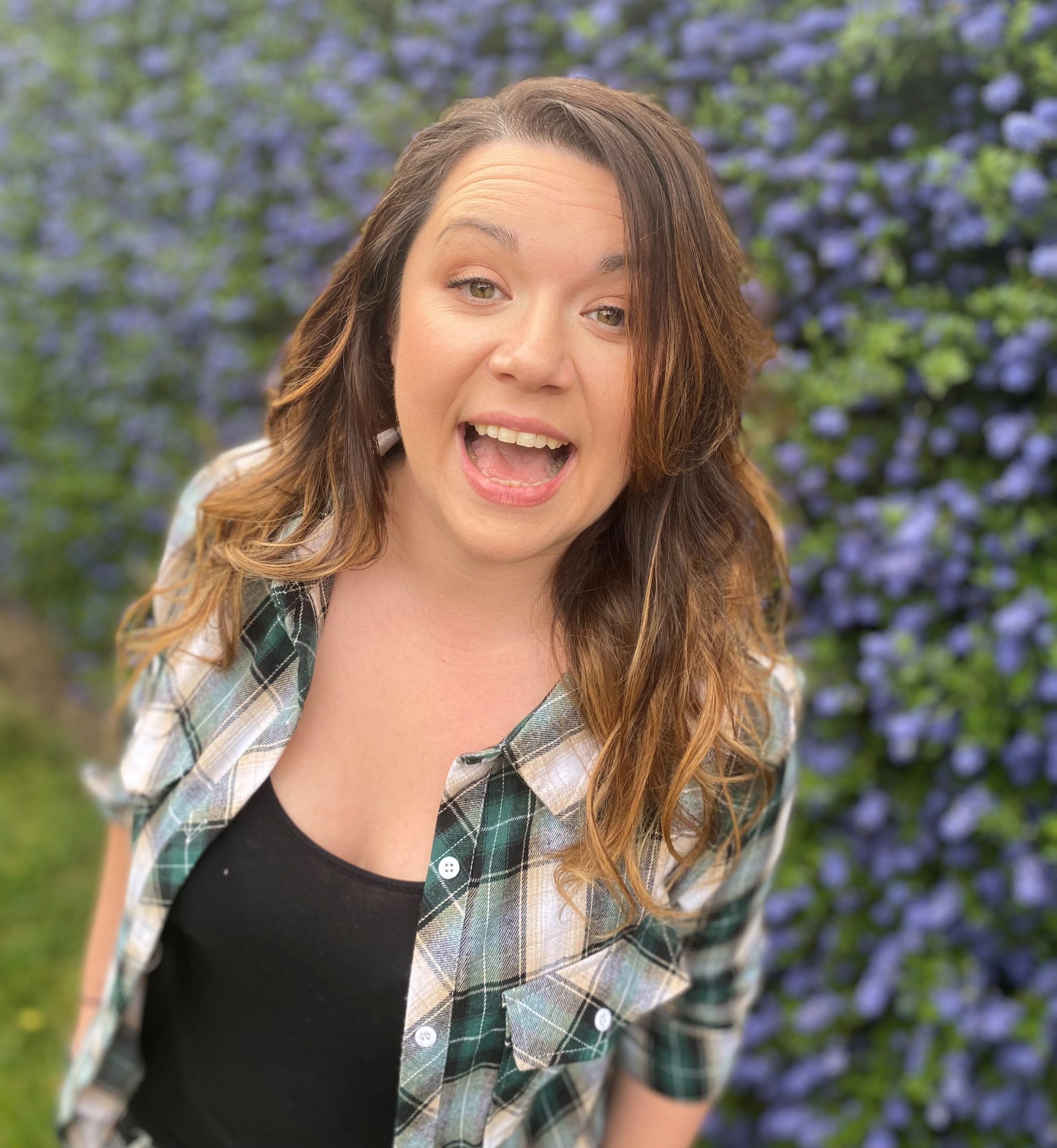 Lizzy, a white woman in her early thirties with long brown hair, smiles a very wide smile. She’s wearing a green check shirt open over a black vest, standing in front of a bush covered in blue flowers.