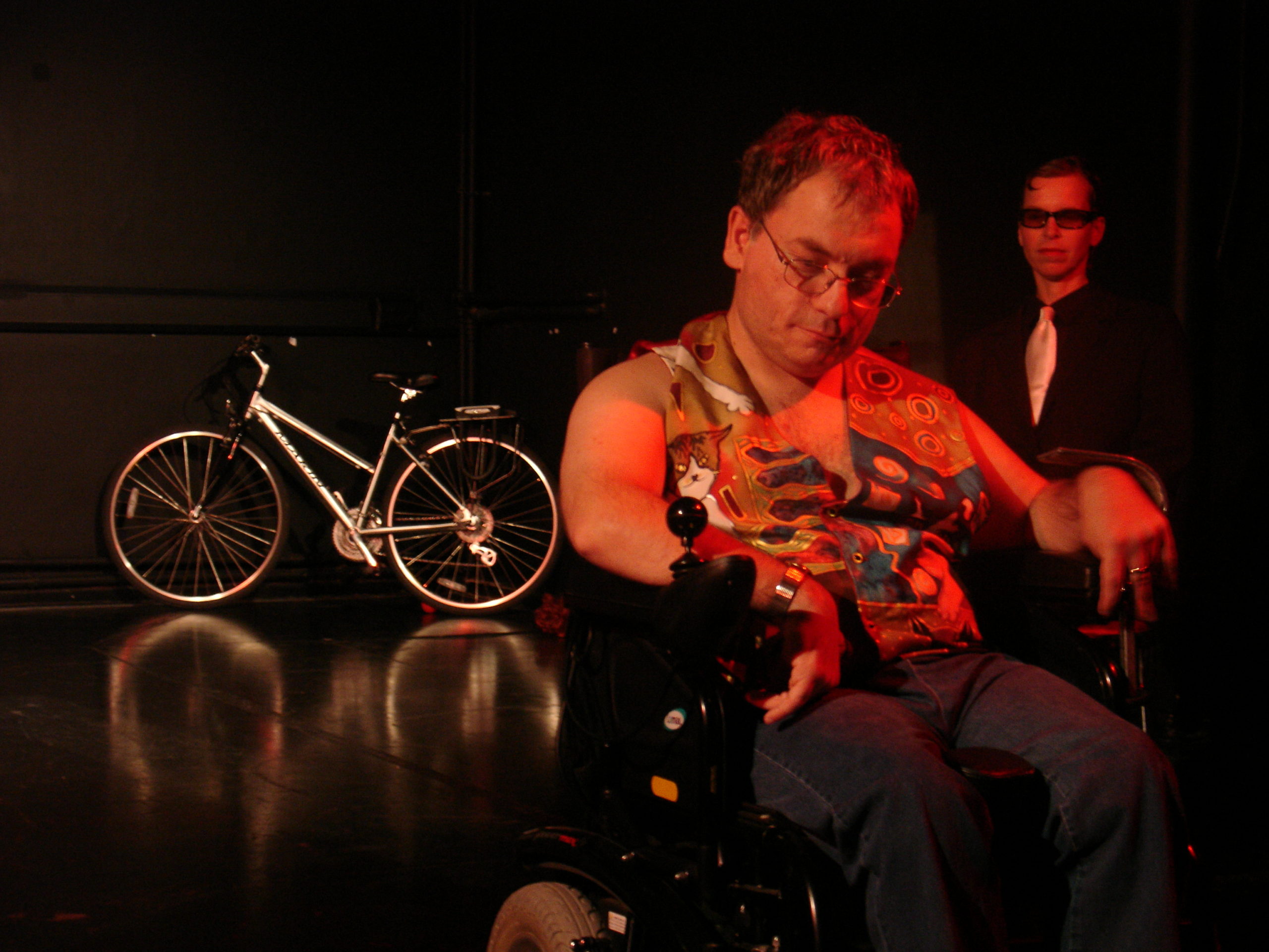 A man in a wheelchair is front and centre stage, he is being watched by a man behind him in sunglasses. There is a bike on the stage behind them.