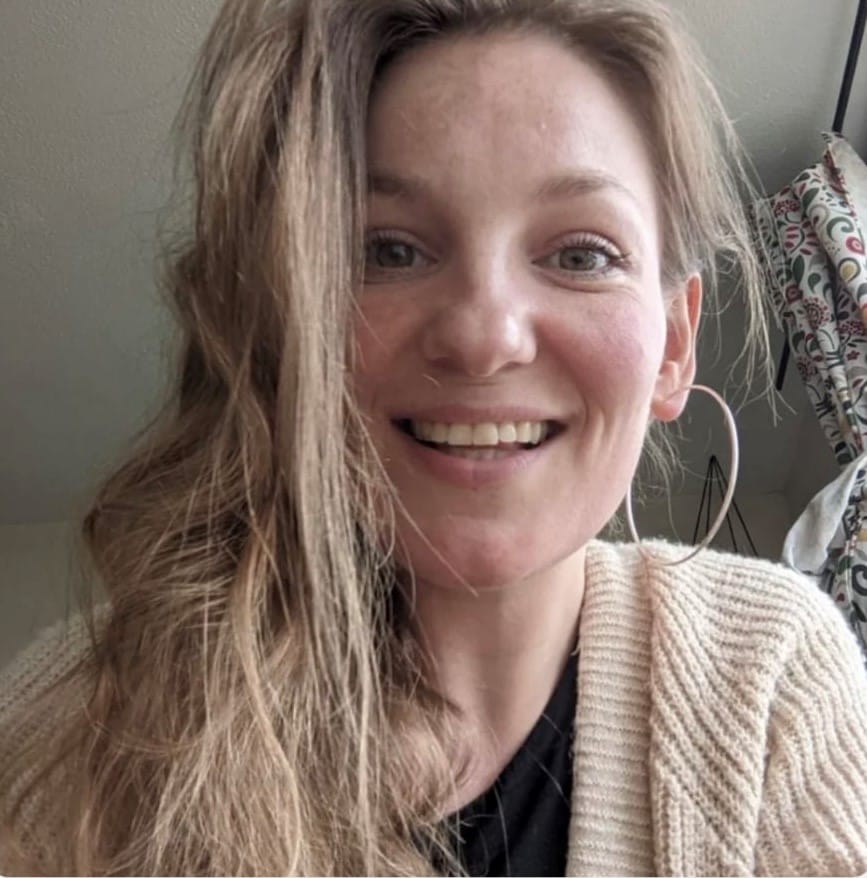 A white woman in her 30s, with long light brown hair swept to the side, wearing very big silver hoop earrings and a big smile