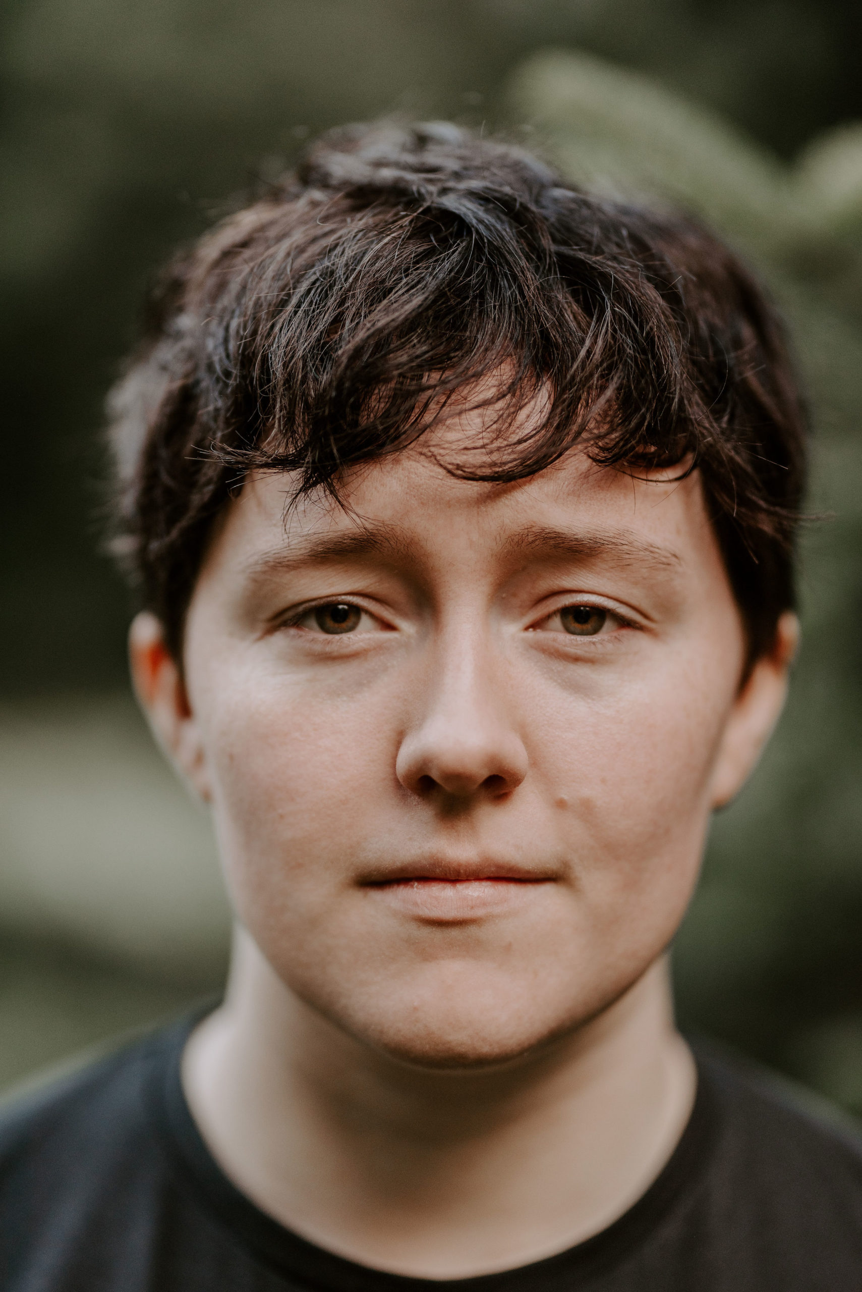 Niamh is a non-binary/trans-masc person in their early 20’s. They are white, with short dark brown hair. They are wearing a plain black t-shirt and stood in front of a blurred green background.