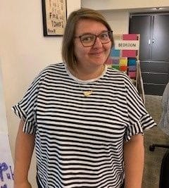 Vicky, a white woman with medium length brown hair and glasses, is stood in our Graeae office. She is wearing a black and white striped t-shirt and a gold necklace. She is smiling.