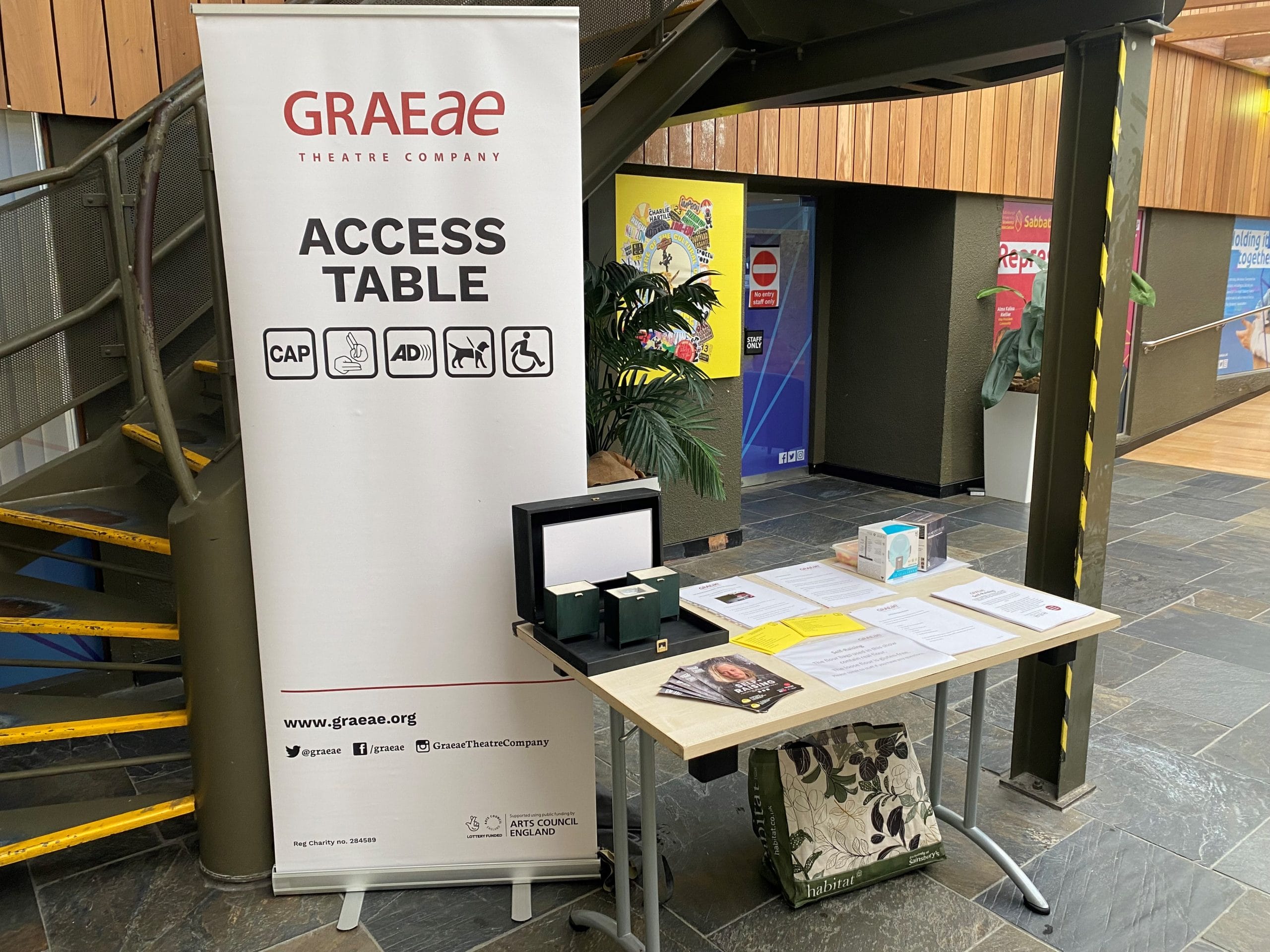 A Graeae access table; there is a large poster to the left of the table that says 'Access Table'. The table itself is littered with flyers and information papers.