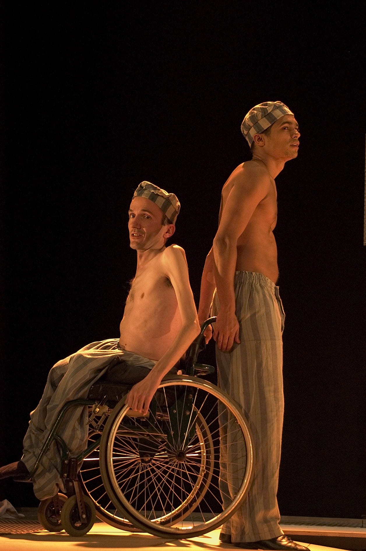 Two performers are positioned back to back, one is in a wheelchair and the other stood upright. They wear the uniform of a person in a concentration camp.