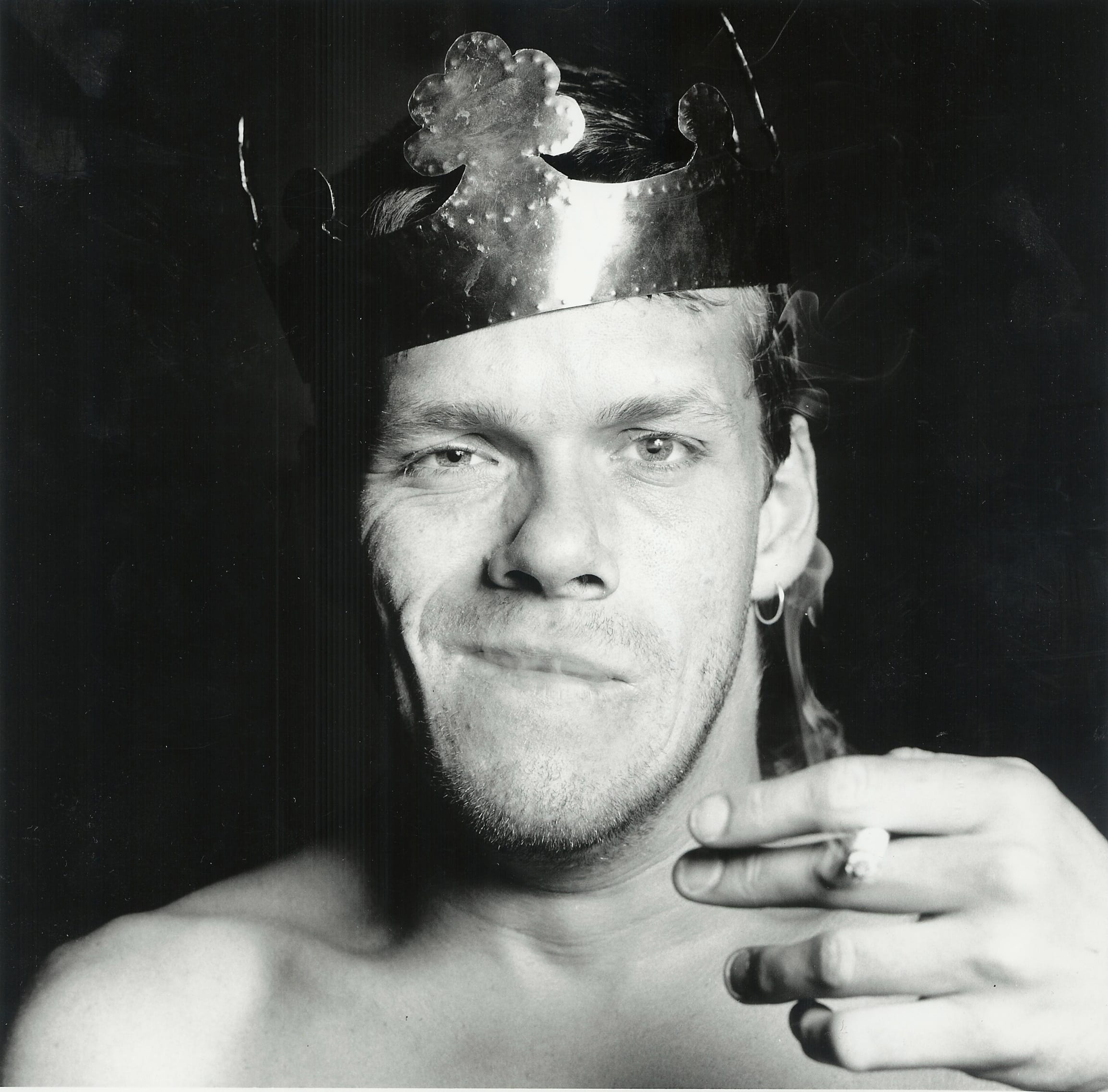 A man faces the camera with a smug expression on his face, he is smoking a lit cigarette and has a crown on his head.
