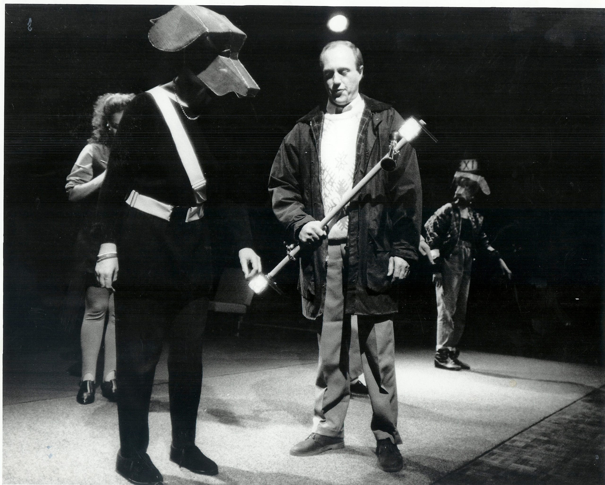 Two performers onstage. One is wearing a large structure on their head, in the shape of a dog. The other is dressed wearing a large woolen jumper and a wax jacket.