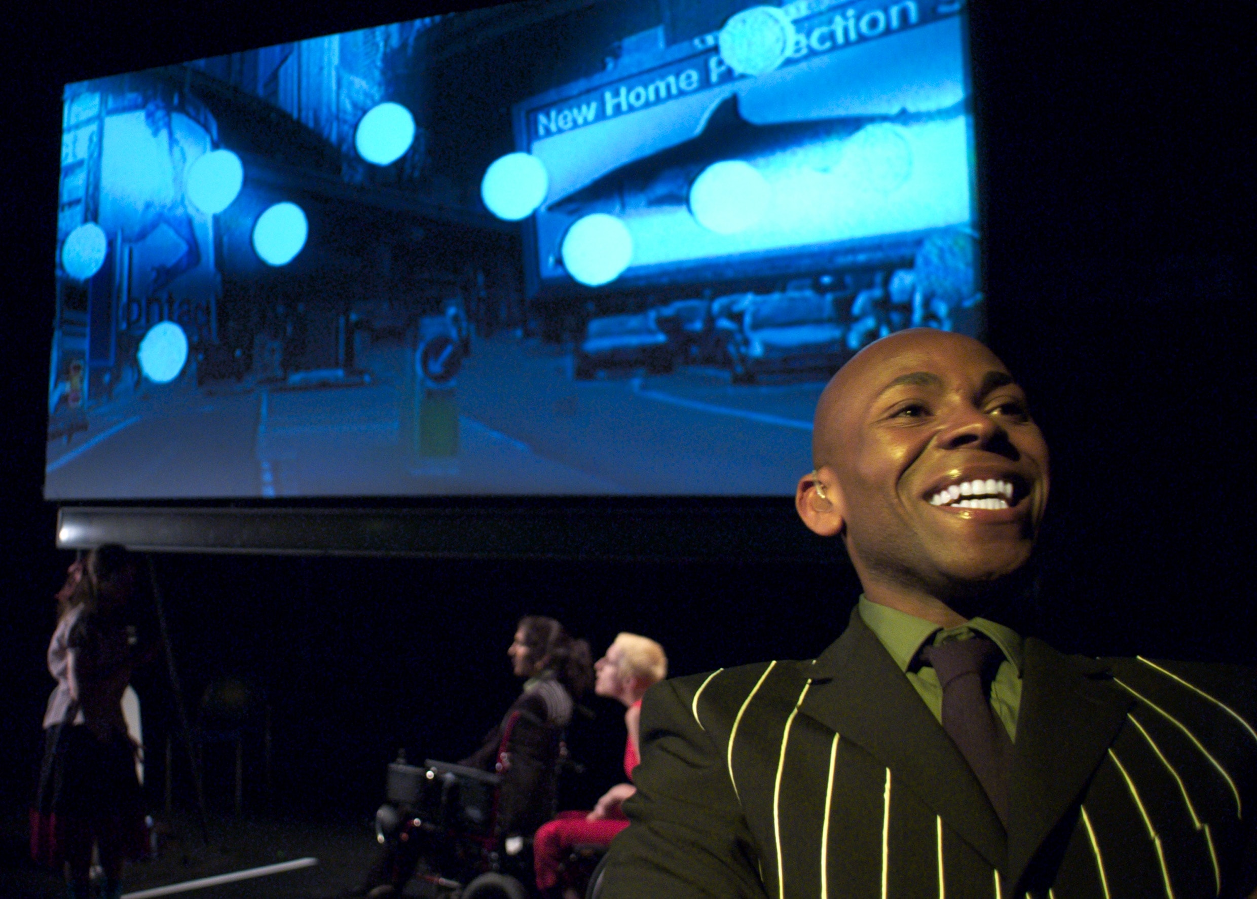 A young black man stands in front of a screen projecting the image of a typical London road. He is beaming a smile.