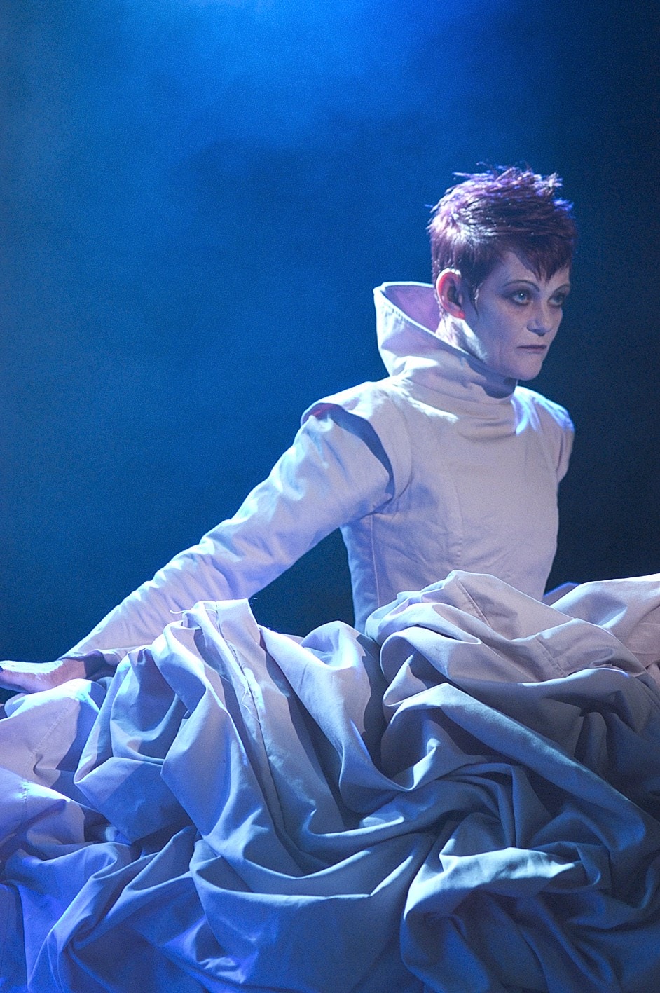 A performer sits amongst swathes of white fabric. They are cast in a blue light.