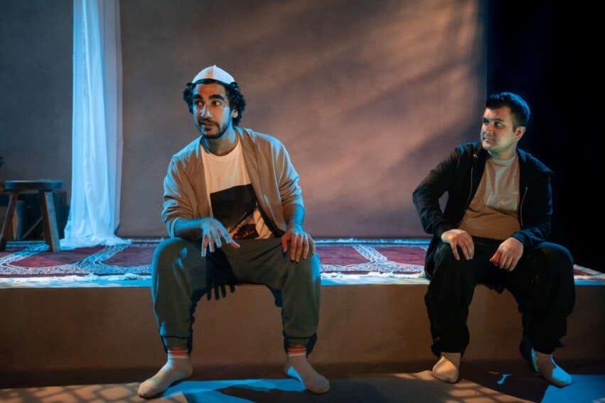 Two male performers sit on the edge of a raised part of the stage. One of them has curly black hair that sticks out from underneath the prayer cap he is wearing. He is speaking and gesturing, as the other man looks across at him and listens. He has short black hair. They are both wearing casual, neutral toned clothes, socks, but no shoes. Their are prayer mats and a stool on the stage behind them.