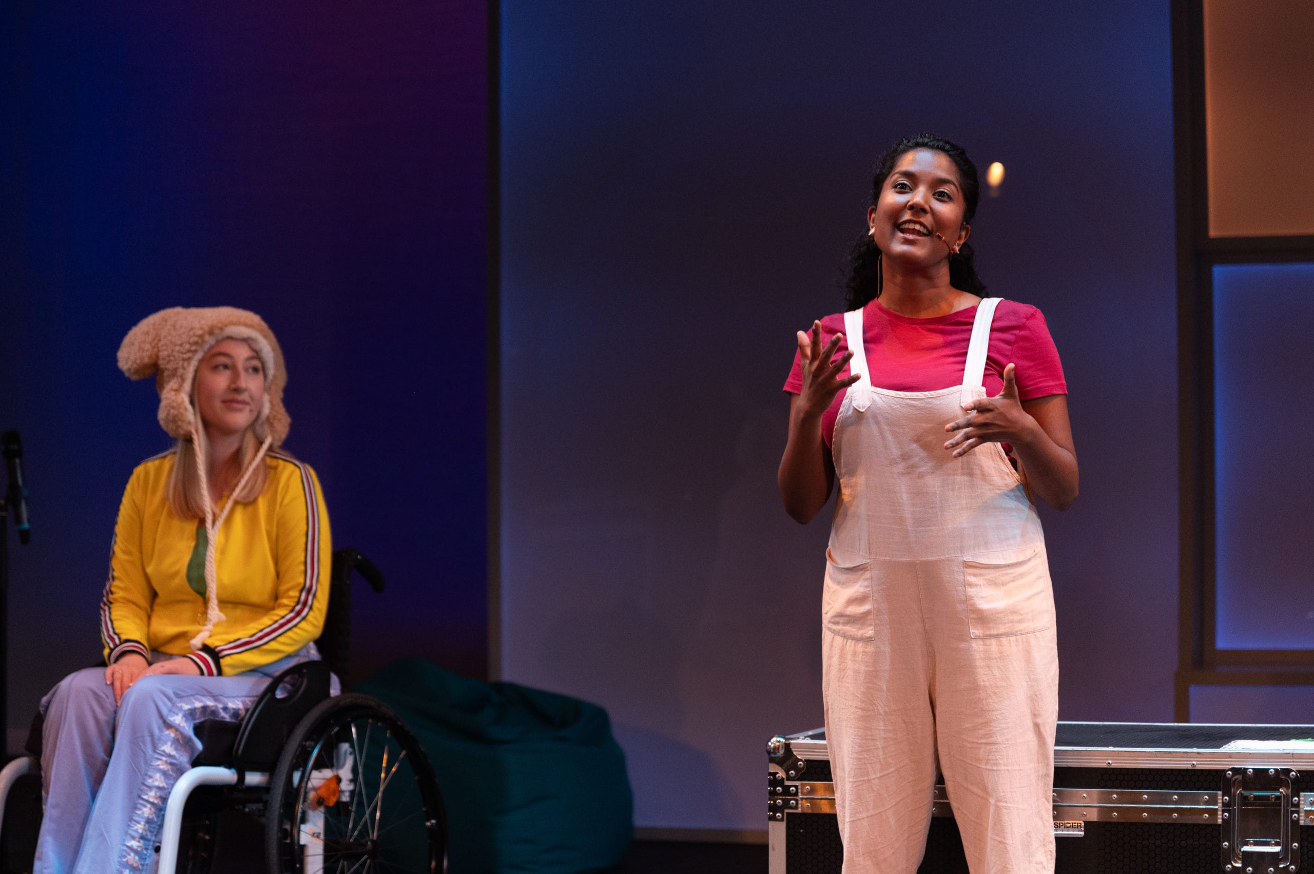 An interpreter performs on stage, signing what she is saying. Another performer in a wheelchair is positioned upstage of her, and is watching her.