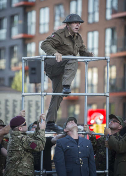 A man dressed in a soldiers uniform and helmet, climbs a piece of scaffolding. Other soldiers are holding up the scaffolding for him. One man stands with his back to the scaffolding and leans backward to look up at the climbing soldier.