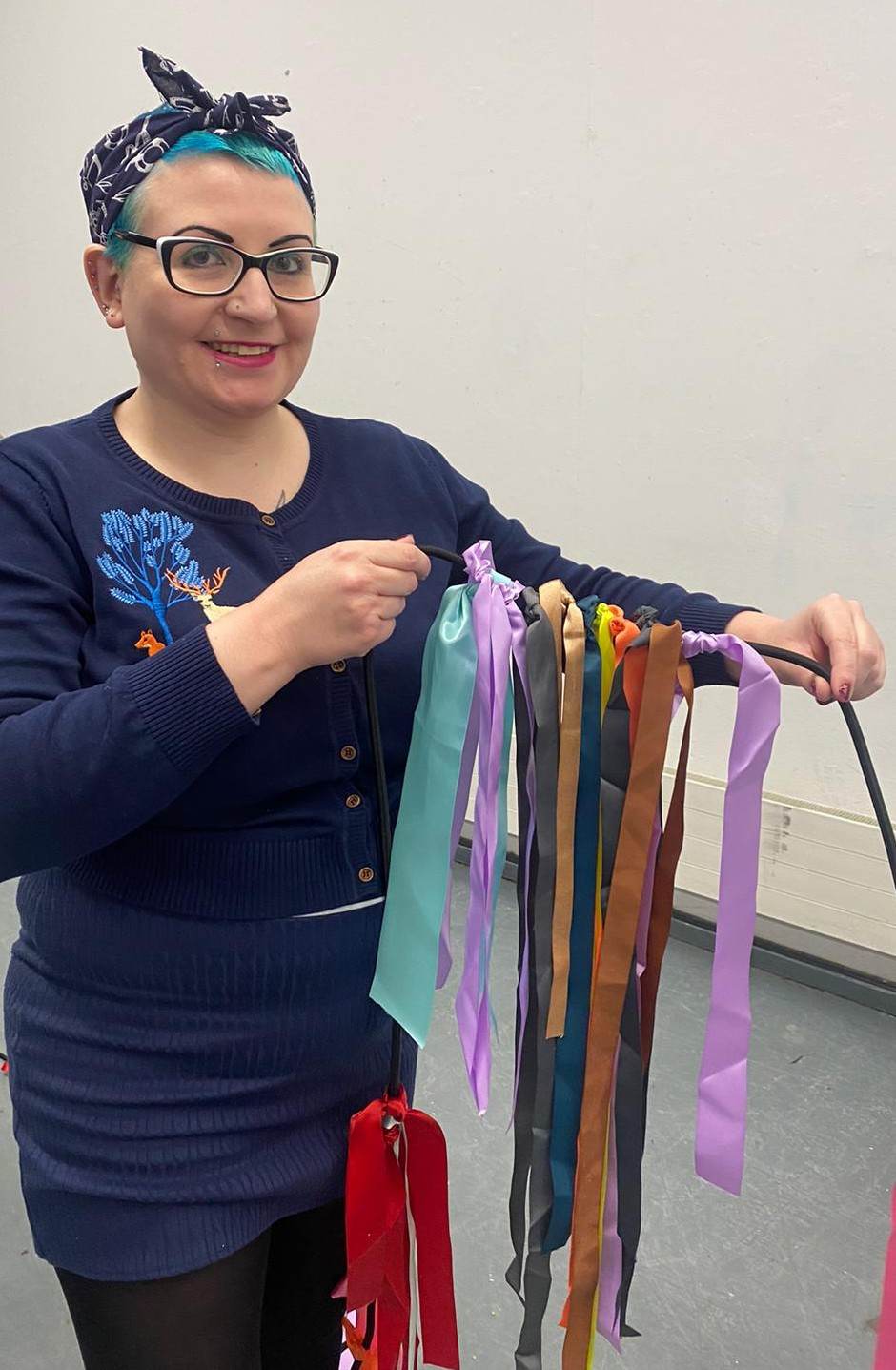 Gemma, a white woman with glasses, and blue hair that she has wrapped in a blue headscarf, holds up a string of ribbons and smiles at the camera.