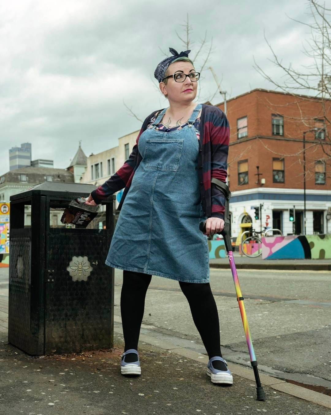 Gemma, a white woman with glasses, and blue hair which she has wrapped in a blue headscarf, stands in the street next to a bin. She is using a walking stick with one hand, and reaching behind her with the other to out something in the bin.