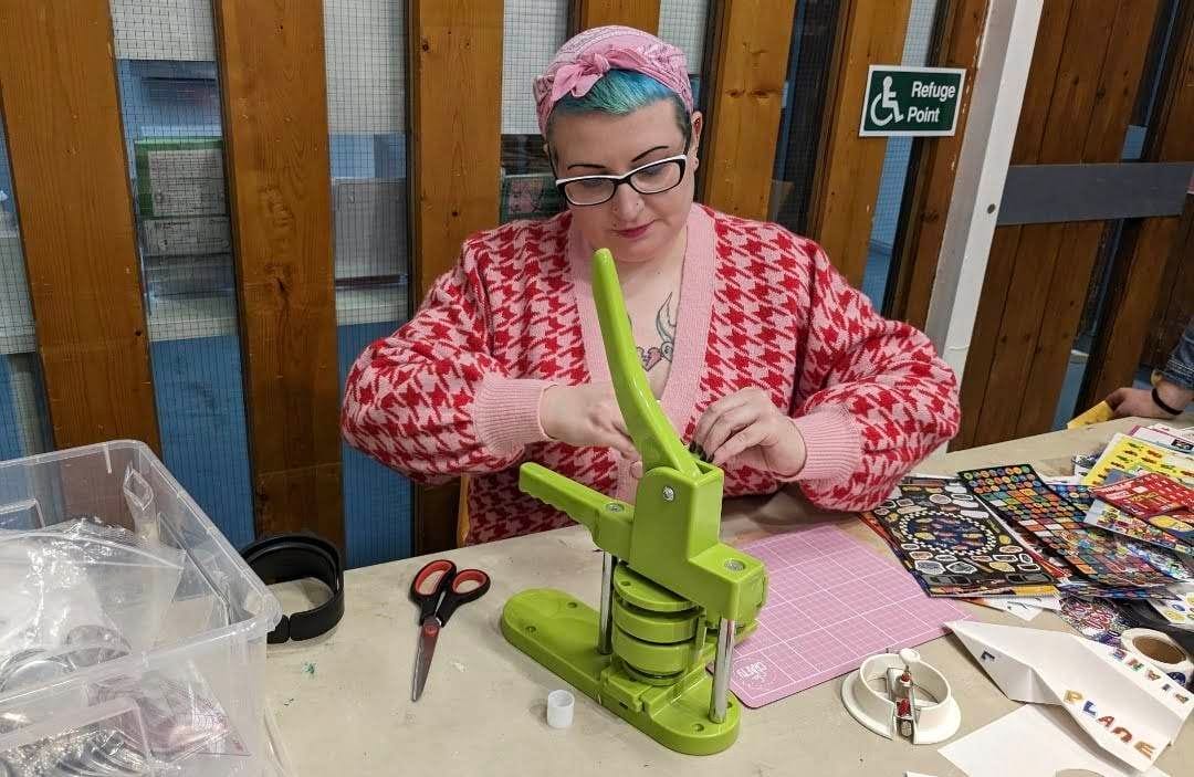 Gemma, a white woman with glasses, and blue hair which she has wrapped in a pink headscarf, uses a green pin badge making machine. There are lots of craft supplies on the table around her.