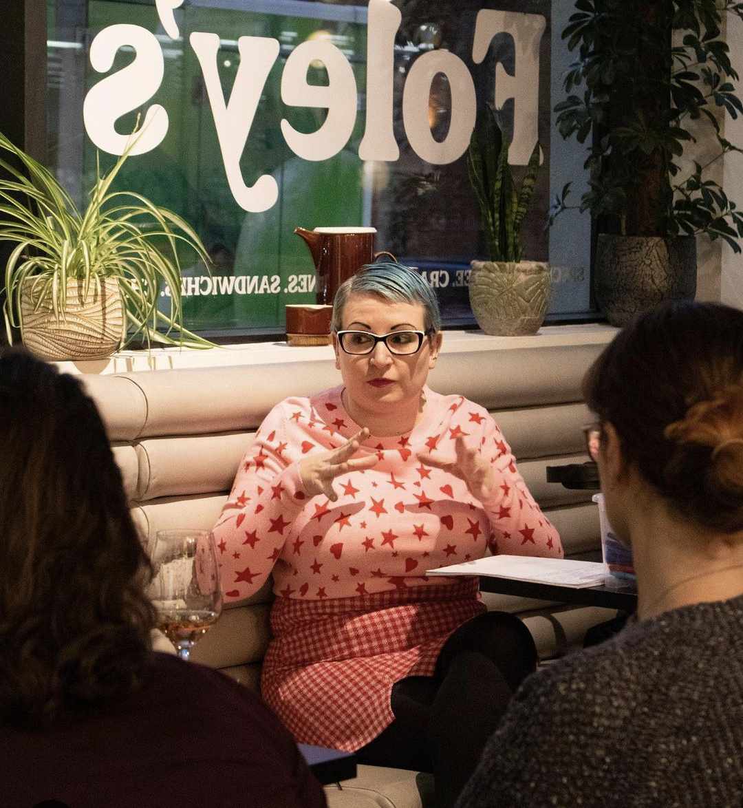Gemma, a white woman with glasses and short blue hair, sits at a table with a few other people. She is wearing a pink jumper with red stars on it, and a pink and red check skirt.